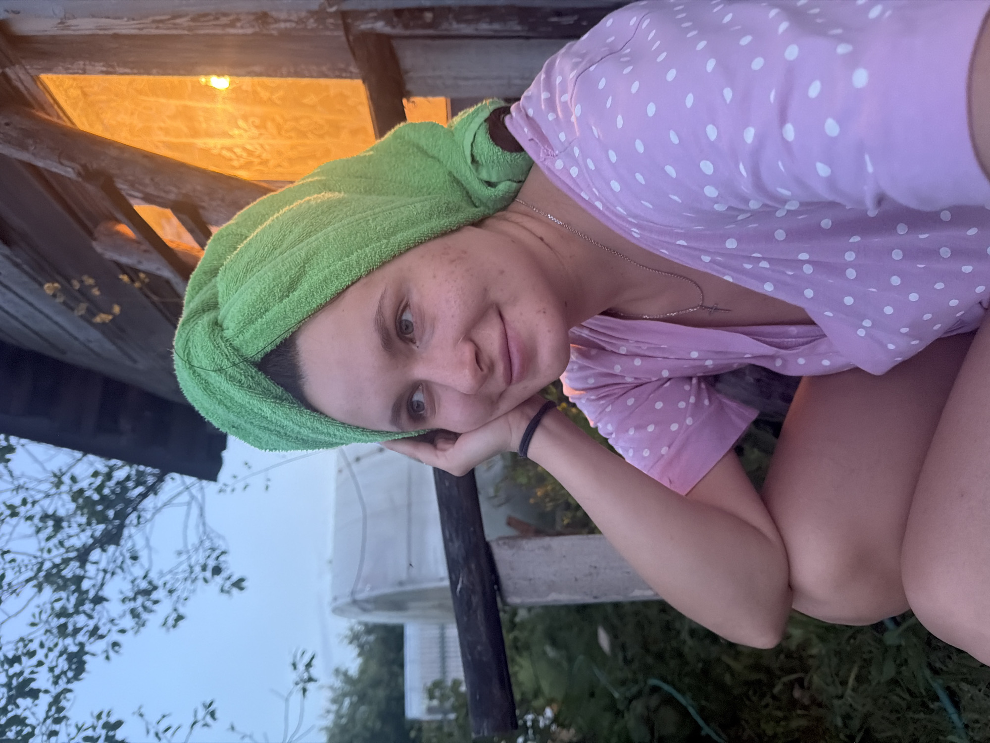 A young woman with freckles and a green towel wrapped around her head sits on a wooden porch in Krasny Mayak, Russia, at dusk. She wears a pink polka-dotted dress and a silver necklace with a cross pendant. Her left arm is bent, and her hand rests on her cheek, supporting her head as she gazes to her left. The background shows a wooden structure with a window emitting an orange glow, trees, and what appears to be a greenhouse or shed. The sky is a muted twilight blue.