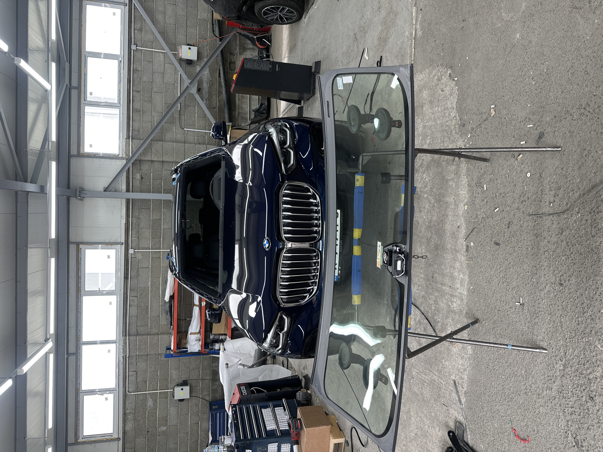In an indoor garage setting, a dark blue BMW X5 is parked, facing forward. In the foreground, a windshield is propped up on a stand, indicating it's either being replaced or has been removed. Tools and equipment are visible around the car and windshield, including a red toolbox and shelves stocked with items. The garage has concrete floors, brick walls, and fluorescent lighting. The overall scene suggests an automotive repair or maintenance operation is in progress.