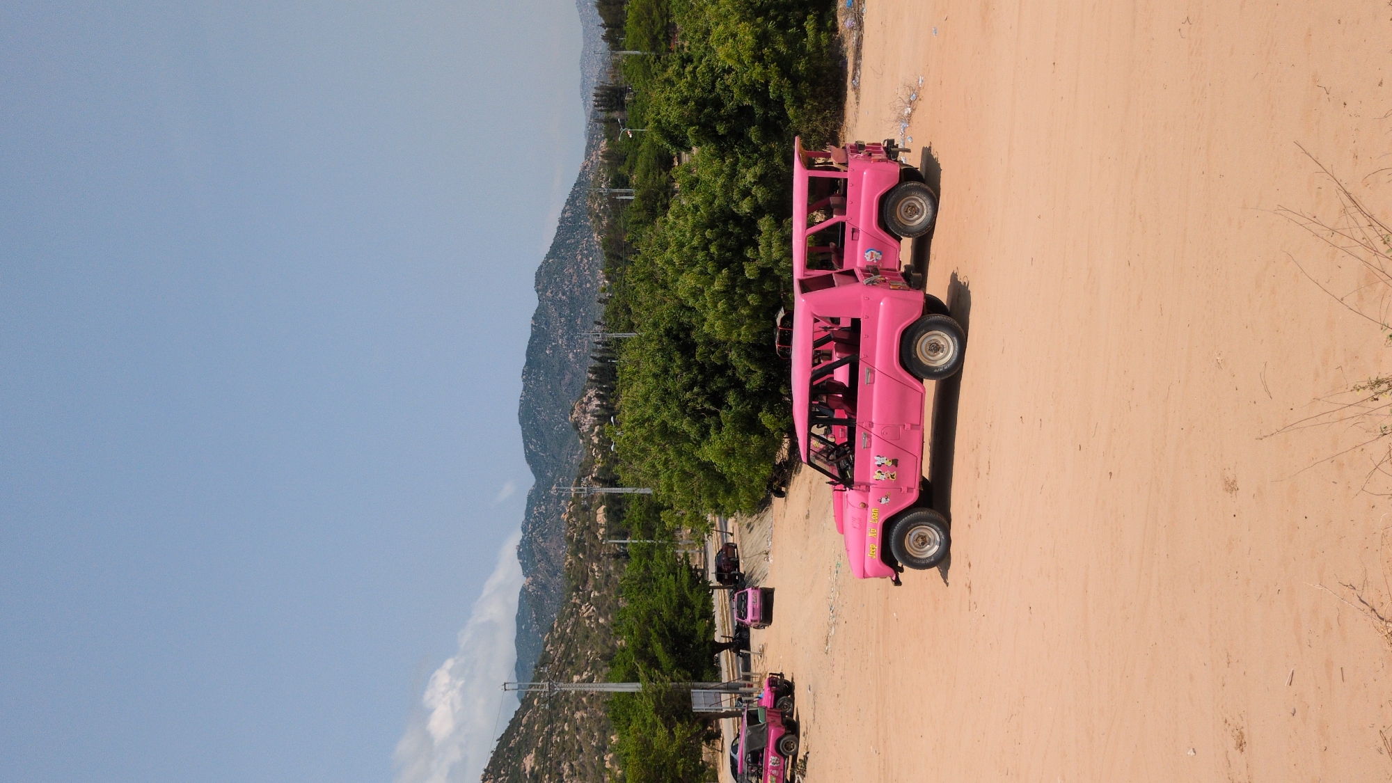 The image depicts a pink Jeep-like vehicle parked on a sandy surface, likely a road or open area. In the background, there are mountains, lush green trees, and utility poles. Additional pink Jeep-like vehicles can be seen farther down the road. The location is likely Phuoc Dinh, Vietnam, based on the prompt.