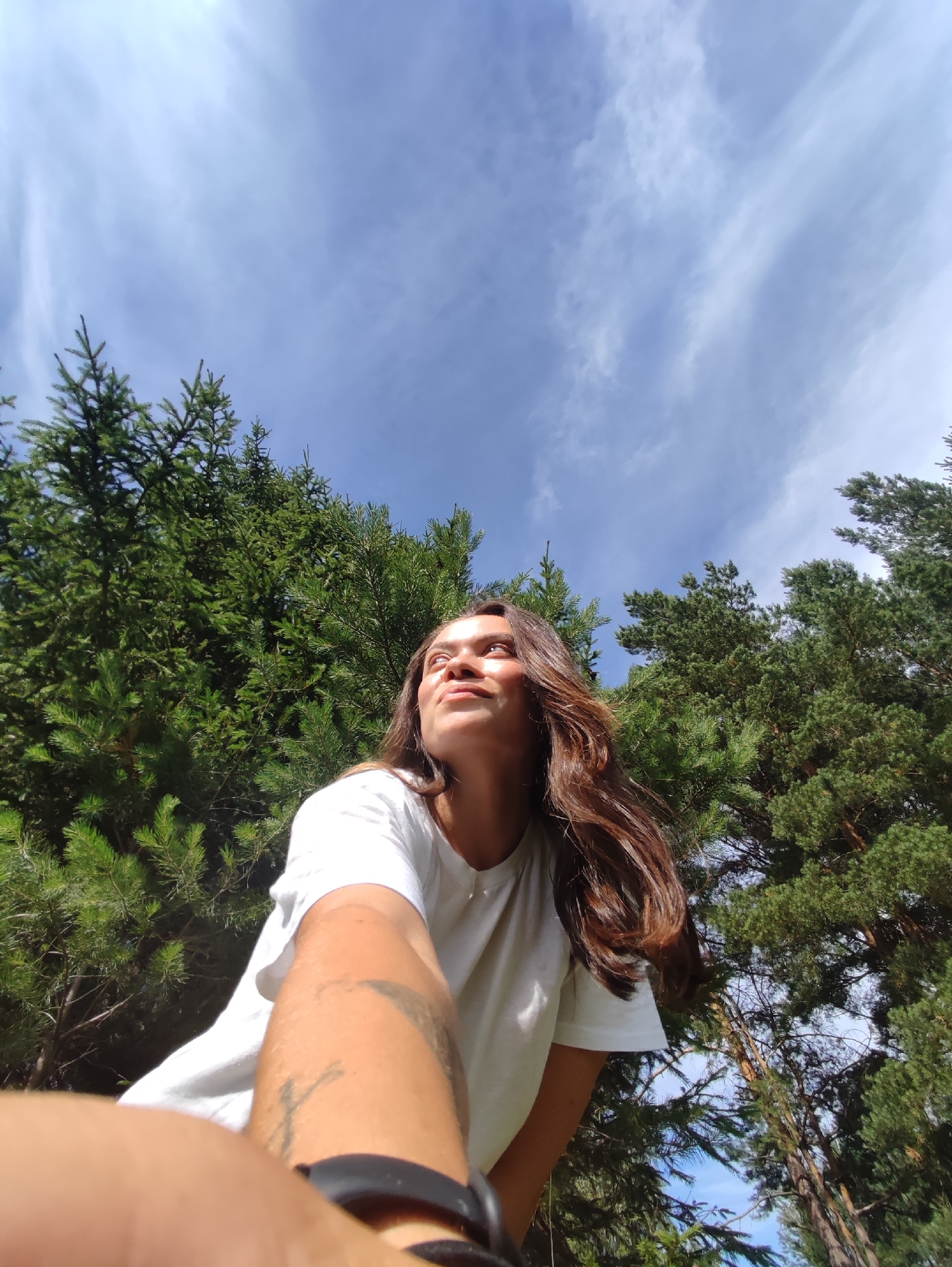 A low-angle, wide shot captures a young woman outdoors, looking up at the sky. She has long, dark brown hair and is wearing a white t-shirt. Her left arm is extended towards the camera, and a black watch is visible on her wrist. Tattoos are present on her left forearm. She is positioned in the foreground, with a dense forest of pine trees filling the background. The sky above is a bright blue with scattered white clouds. The location is the city of Фоки, Russia.