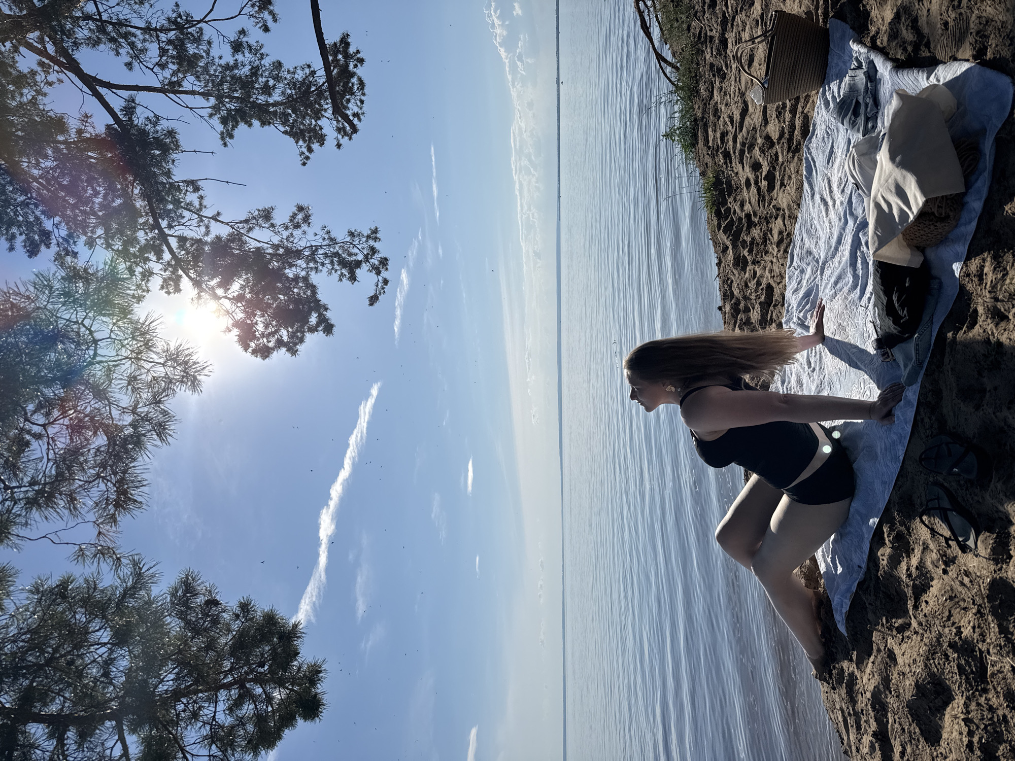 A woman sits on a towel on a sandy shore, facing the water, with her head tilted back and eyes closed, basking in the sun. She is wearing a black swimsuit. A beach bag and some clothing are laid out on the towel beside her. The sun shines brightly in the sky, partially obscured by pine tree branches overhead. The calm water stretches to the horizon under a blue sky with scattered clouds and many small birds flying.