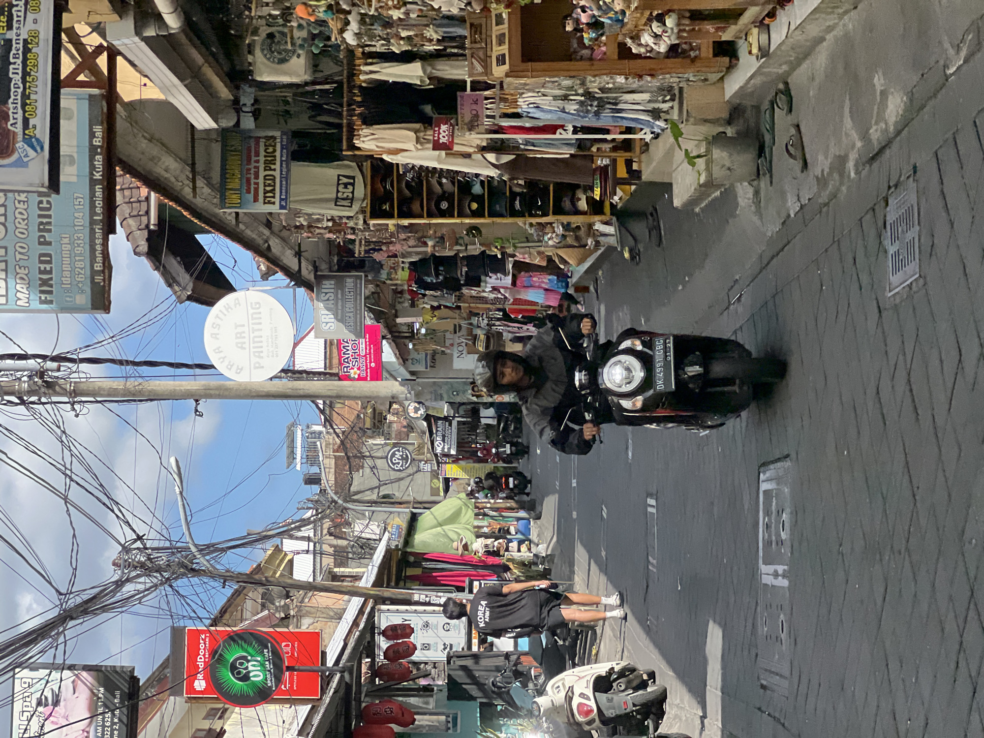 The image depicts a narrow street in Kuta, Bali, Indonesia, lined with shops on both sides. The street is paved with interlocking bricks. Overhead, a dense network of electrical wires is visible.
In the foreground, a person wearing a helmet and a dark jacket is riding a black scooter. The scooter has a license plate that reads "DK 4991 GBG". To the left of the scooter rider, a person wearing a black t-shirt with "KOREA ARMY" printed on it and black shorts is walking away from the camera. Several scooters are parked on the left side of the street.
The shops on the right side of the street display various goods, including clothing, souvenirs, and accessories. Some shops have signs with text such as "TONY MACARONY SHOP" and "FIXED PRICES". On the left side of the street, there are shops with signs like "RedDoorz" and "UNI SPORT BAR CAFE".
The sky above is partly cloudy. The overall scene suggests a bustling commercial street in a tropical urban environment.