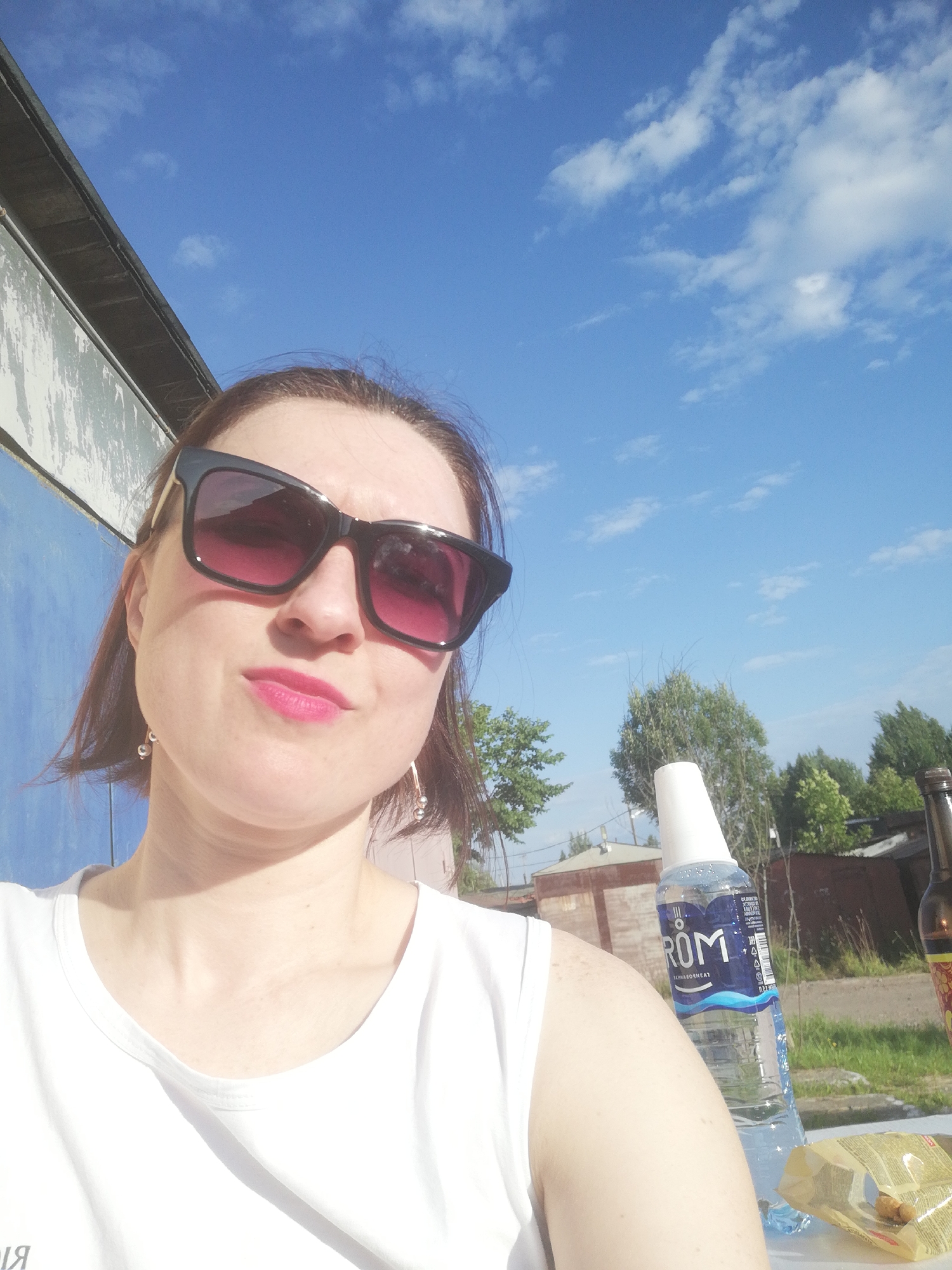 A close-up, selfie-style portrait of a woman with brown hair, dark sunglasses, and bright pink lipstick, wearing a white t-shirt. She is outdoors with a blue sky and scattered clouds in the background. To her right, a plastic bottle of "ROM" water and a partial view of a brown bottle are visible on a table. The background suggests an urban or semi-urban setting with buildings and trees. The overall scene appears to be taken during daylight, possibly in the city of Tosno, Russia, given the context of the request.