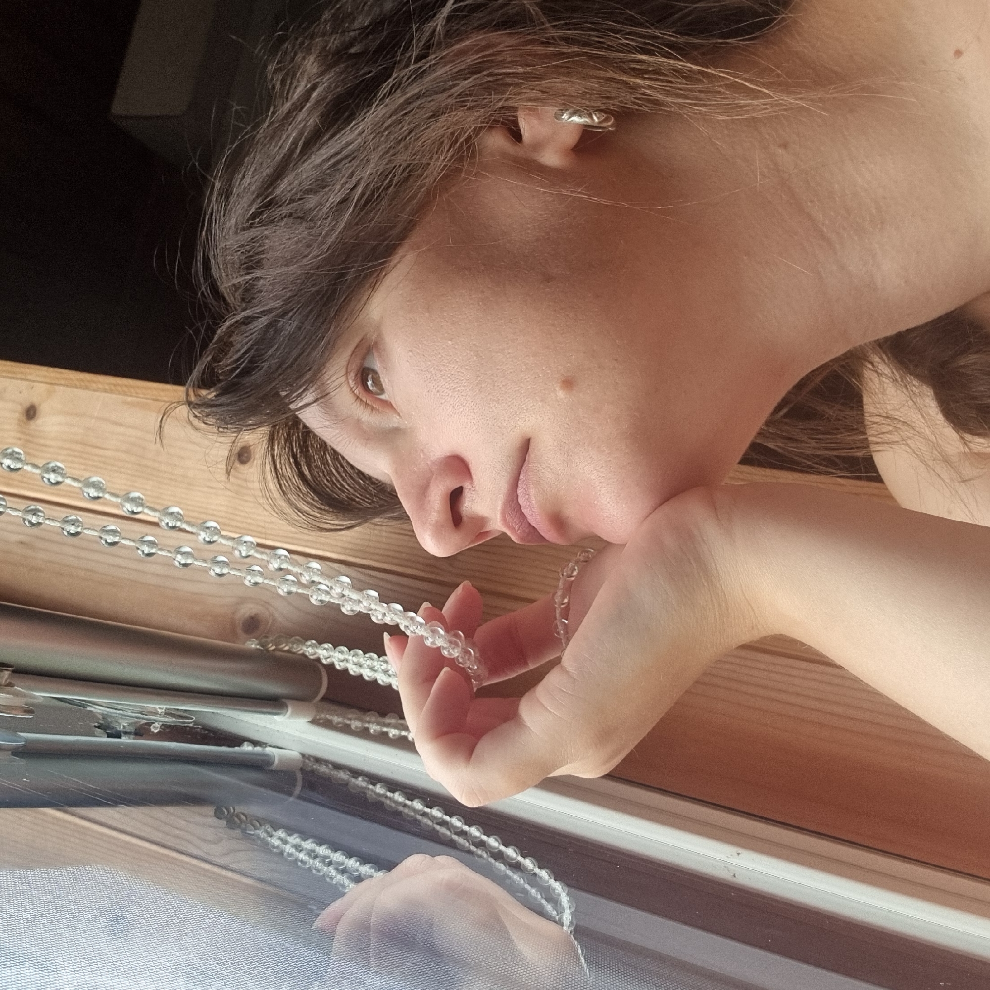 An extreme close-up, side profile shot shows a young adult female, with dark brown hair and fair skin, gazing upwards and to the right out of a window. She is resting her chin on her hand, which is holding onto a strand of clear beaded garland. The garland hangs vertically in front of her face and is reflected in the window. Her left earlobe, adorned with a silver hoop earring, is visible. The background consists of a wooden surface and the window frame. The environment appears to be indoors, possibly a home, with natural light illuminating the scene. The location of Dunnae-myeon, South Korea is not depicted in this image.