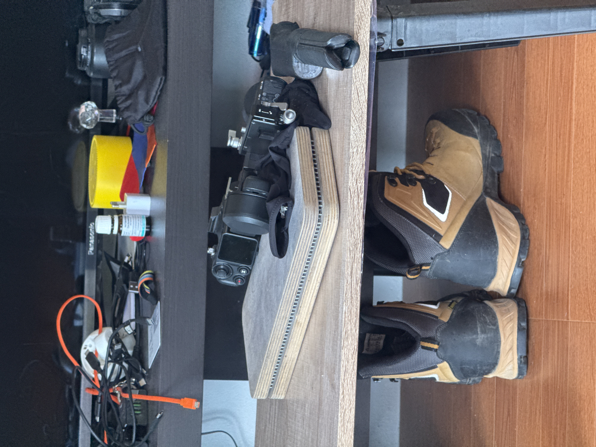 The image depicts an indoor scene, likely a living room or storage area, on the island of Ko Samui in Thailand. The room is sparsely furnished, with items organized on shelves. The top shelf displays a tangle of wires, including an orange ethernet cable, a white device, and a small bottle of essential oil. A yellow cylindrical object, possibly a speaker, and a black fabric pouch are also visible on this shelf.
The middle shelf holds photographic equipment. A camera with a lens is positioned on a flat wooden platform. Nearby, there are black cloth accessories and a black cylindrical object, possibly a lens cap or grip. A blue object, possibly a flash or other accessory, is also present on this shelf.
The bottom shelf displays a pair of brown and black hiking boots, suggesting outdoor activities are a part of the owner's lifestyle. The floor is a warm-toned wooden laminate. The overall impression is one of organized clutter, with a focus on electronics and outdoor gear. There are no people visible in the image.