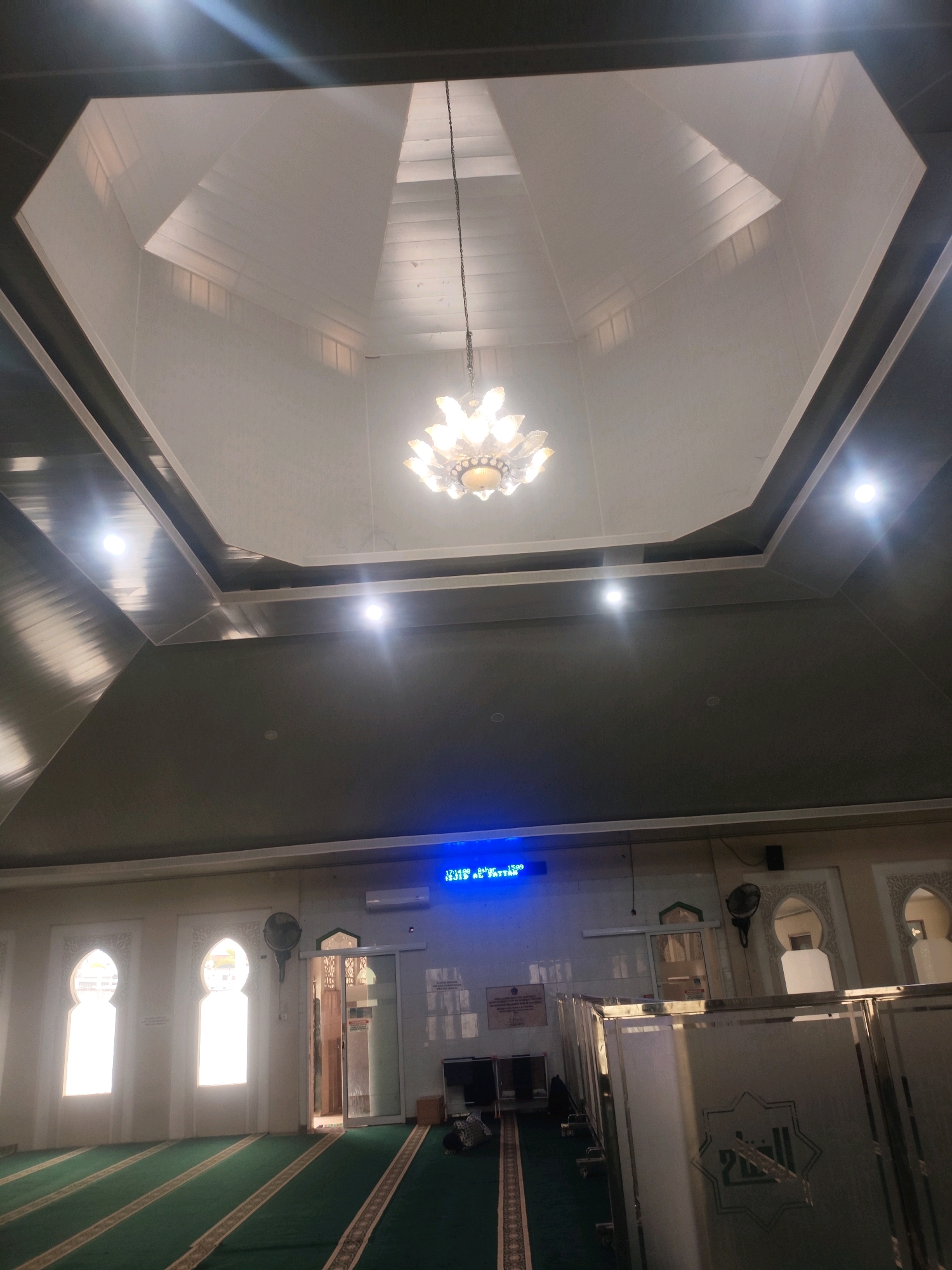 The image shows the interior of a mosque with a hexagonal-shaped ceiling. A modern chandelier hangs from the center of the ceiling. The walls are painted white and adorned with decorative archways. Large windows with intricate patterns allow natural light to enter the prayer hall. The floor is covered with green carpet marked with yellow stripes. In the background, a digital clock displays the time and prayer schedule. The mosque appears to be clean and well-maintained, with no visible signs of damage or disrepair. The overall impression is one of tranquility and reverence, characteristic of a place of worship.