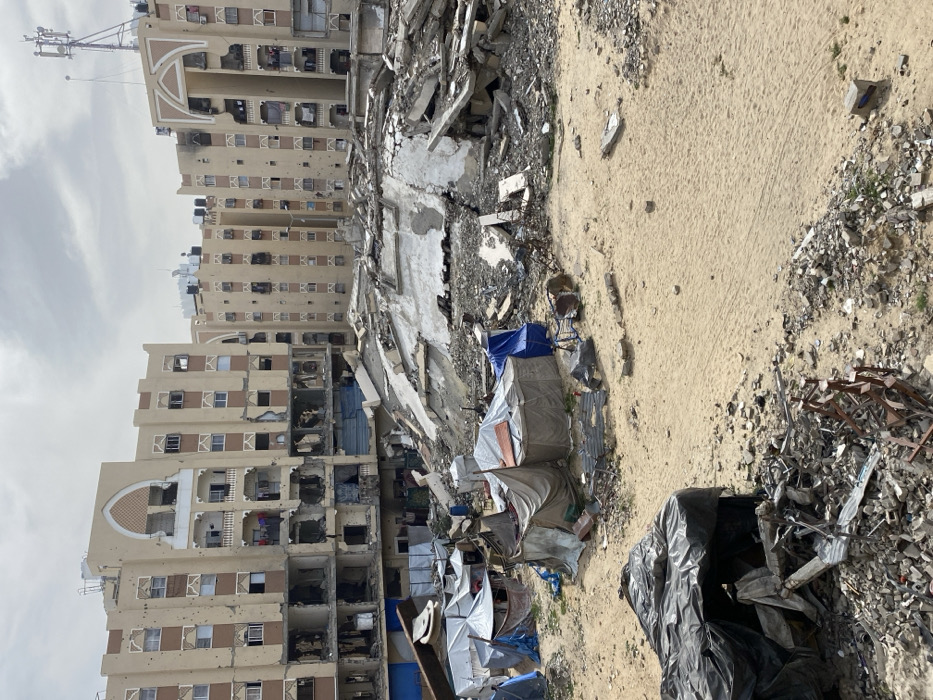 The image depicts a severely damaged urban landscape in Khan Yunis, Palestinian Territory, under an overcast sky. In the background, multiple multi-story buildings, approximately seven to eight floors high, exhibit extensive structural damage. These light-colored buildings, featuring distinctive arched window frames and balconies, are pockmarked with numerous impact holes. Lower floors show significant collapse, with large sections of exterior walls missing, exposing interiors and twisted rebar. Water tanks are visible on rooftops, and a telecommunications tower stands atop the building on the far right. Laundry hangs on a balcony railing on one of the partially damaged buildings.
In the midground, a substantial mound of concrete rubble, rebar, and shattered masonry represents the remains of at least one completely collapsed structure, forming a large debris field.
The foreground and midground consist of a sandy, uneven terrain interspersed with smaller debris and refuse. Scattered across this area are numerous makeshift shelters or tents, constructed from various materials including blue, grey, and black tarpaulins, fabric, and salvaged metal sheets. These improvised structures vary in size and shape. A large structure covered in black tarpaulin occupies the bottom left amidst smaller rubble. No discernible individuals are visible, though the presence of tents and laundry indicates human activity and habitation.
