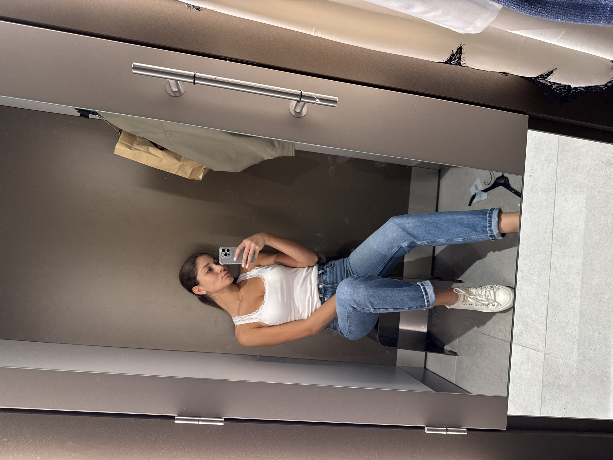 A young woman is captured in a mirror selfie, taken inside what appears to be a fitting room, likely in Burgas, Bulgaria. She is positioned on her side, leaning against a dark wall, while holding a smartphone with her right hand to take the photo. Her head is slightly tilted upwards, and she looks directly at the phone's camera, displaying a neutral expression. She has long brown hair, pulled back, and wears a white, lace-trimmed tank top, blue denim jeans cuffed at the ankles, and white lace-up sneakers. Small stud earrings are visible, and her fingernails are painted white.
The setting is an indoor space, strongly suggesting a retail fitting room due to the presence of a large, full-length mirror, a metal clothing rail, and what appears to be a curtain or fabric at the top of the frame. Above the woman's reflection, a light-colored garment and a brown paper bag hang from the metal rail. A black clothes hanger is also visible in the mirror's reflection to the right of the woman's legs. The floor visible in the mirror is covered with light-colored square tiles. The wall behind her reflection is a plain, dark brown or grey. The lighting is artificial and consistent with an indoor environment. No discernible text is visible on any objects or surfaces in the image.