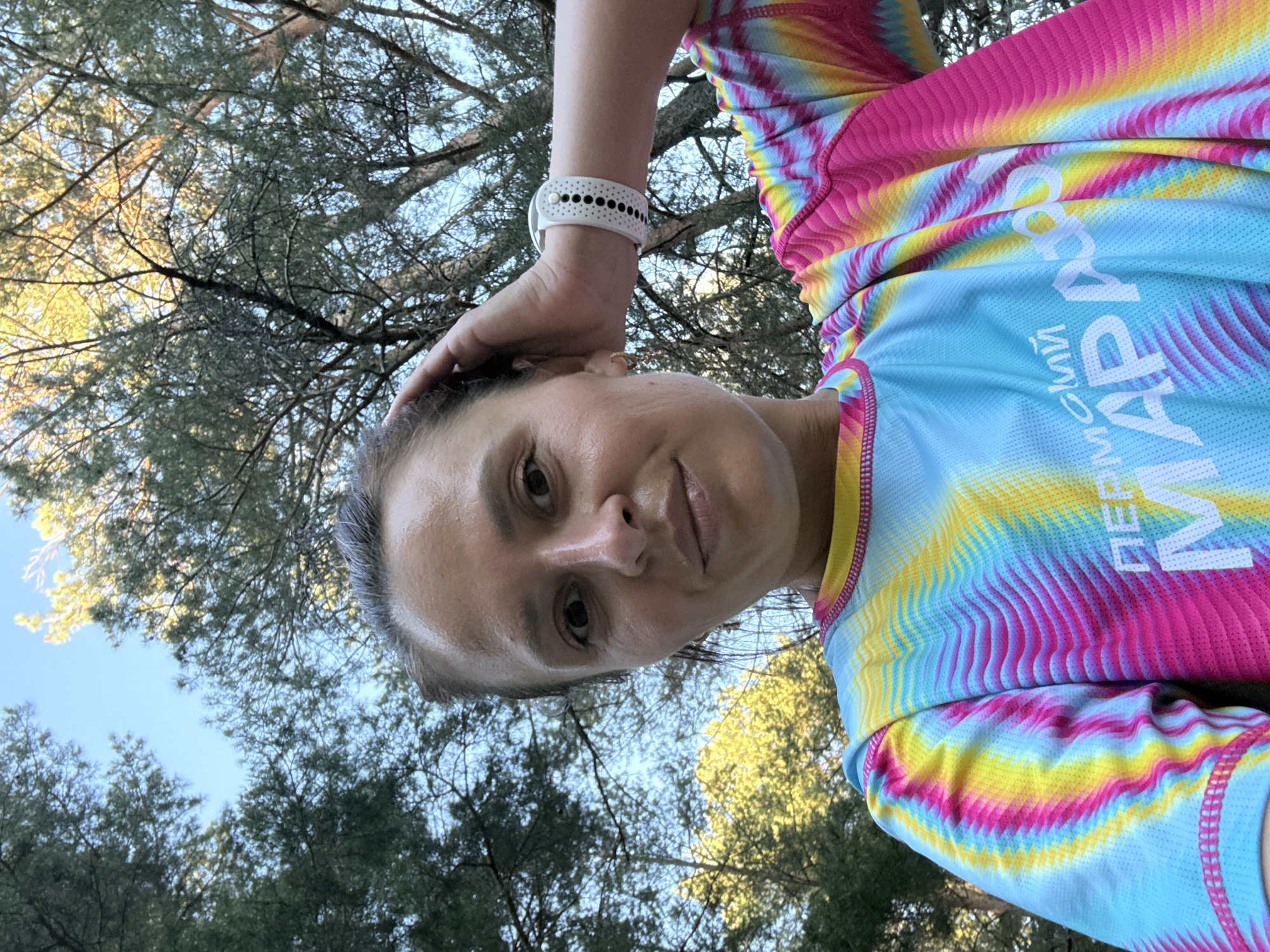 A selfie of a woman in Chaikovsky, Russia. She is wearing a colorful running shirt with the word "ПЕРМСКИЙ" visible on it, a white watch, and she has one hand on her head. The background shows a tree canopy and a glimpse of the sky.