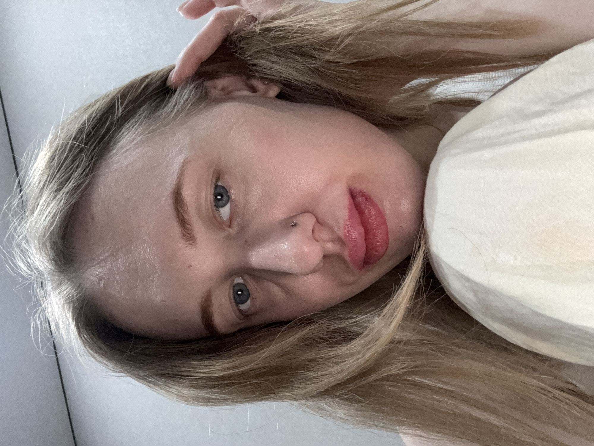 The image presents a close-up indoor portrait of a female individual, identified as being in Rostov-on-Don, Russia. The subject possesses long, light-colored hair and blue irises, maintaining direct eye contact. A small metallic stud is visible on the left side of the nose. The lips are full, exhibiting a reddish-pink hue. The left hand is positioned with fingers near the left temporal region, interacting with the hair. Garments visible include a light-colored top and a cream-colored fabric item in the lower foreground. The background consists of an unadorned light-toned wall.