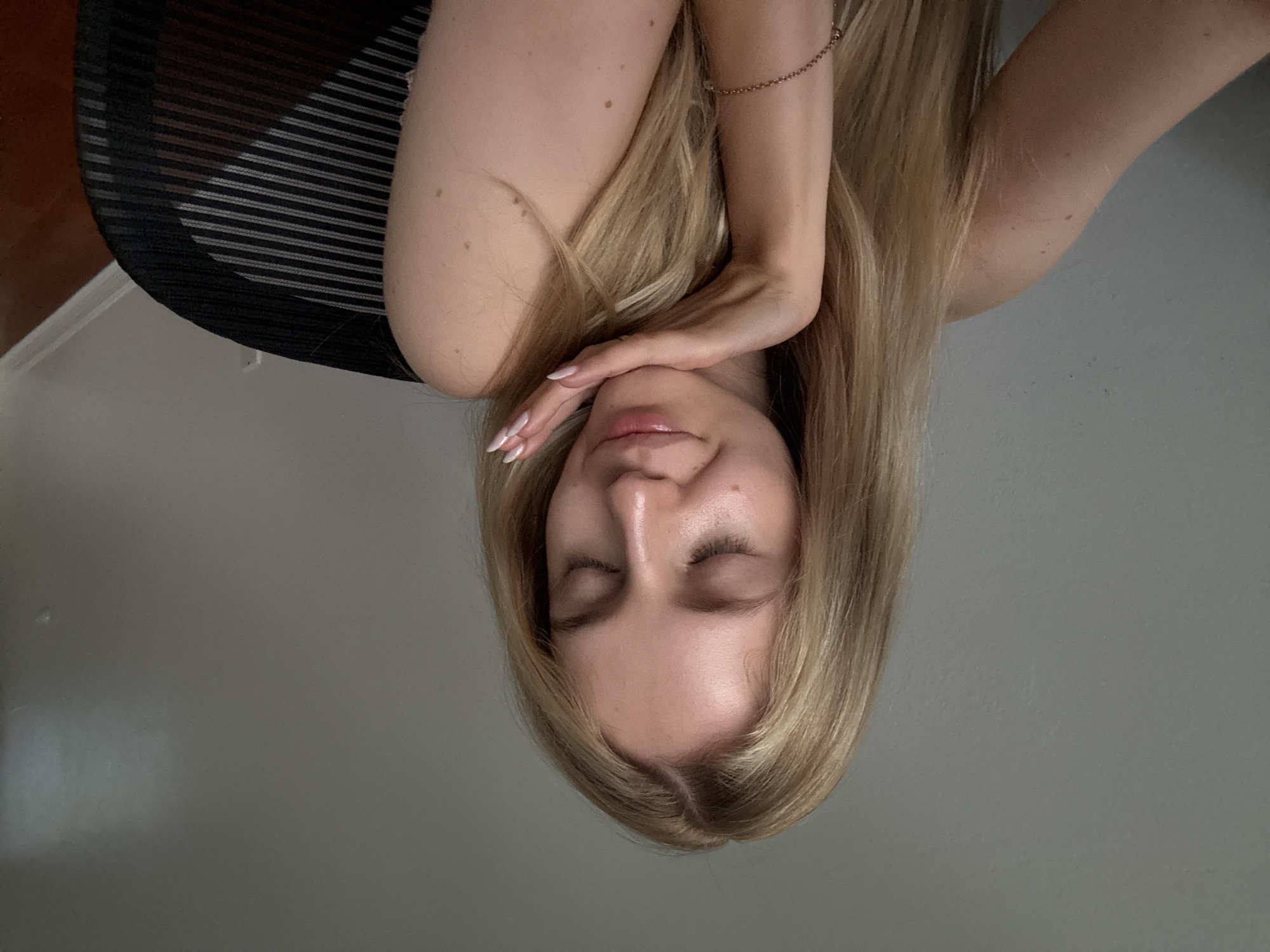 In a medium shot, a young Caucasian woman with blonde hair and closed eyes is seated in front of a plain grey wall. Her left hand is raised, with her palm supporting her chin and her fingers resting near her cheek. She is wearing a light-colored top and a thin bracelet on her left wrist. To her right, a portion of a black office chair with a mesh back is visible. The background suggests an indoor setting, potentially an office or home. No specific city or country can be determined from the visual cues.