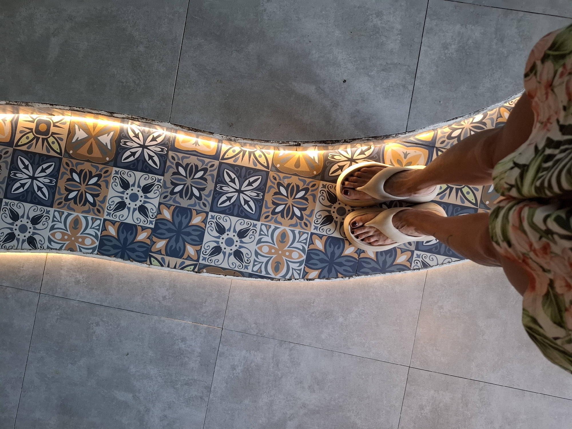 In Nha Trang Ward, Vietnam, a person stands on a curved pathway. The pathway is adorned with colorful, patterned tiles featuring floral designs in shades of blue, yellow, orange, and white. The pathway is bordered by an illuminated strip of light. To the left of the pathway are large, gray floor tiles. The person is wearing light-colored flip-flops and a floral-patterned skirt. The image is taken from a high-angle perspective, looking down at the person's feet and the surrounding floor.