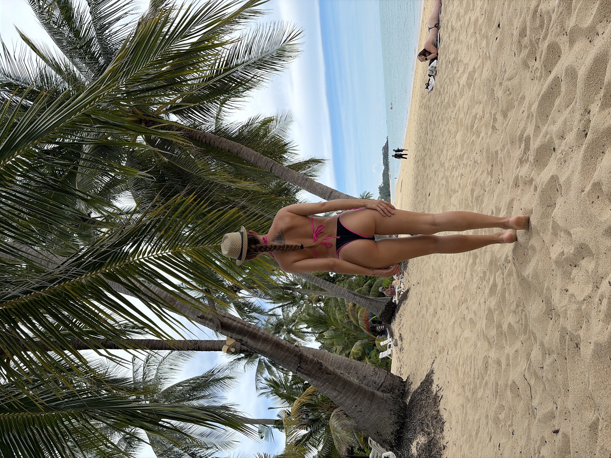 A rear view of a woman wearing a straw hat and a black and pink bikini stands on a sandy beach in Baan Mae Nam, Thailand. She has a braid extending down her back. She is positioned under the fronds of a palm tree, with more palm trees and lush vegetation visible behind her. In the background, the ocean meets a cloudy sky. Several lounge chairs are scattered on the sand, and a few people can be seen walking along the shoreline in the distance. To the right, another woman is lying on a towel on the beach.