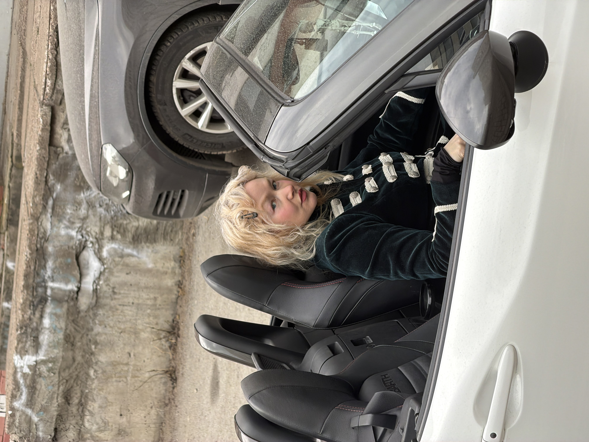The image shows a woman with fair skin and blonde curly hair sitting in the driver's seat of a white convertible car. She is wearing a dark green velvet jacket with light-colored decorative details. Her face is turned towards the camera. The car is parked next to a grey sedan. The background shows a rough concrete surface, possibly a street or parking area.
Based on the Cyrillic text "ЗАРТИ" visible on the passenger seat, it's likely the word "Земля" (Zemlya), which means "Earth."
The image lacks definitive landmarks, but elements like the concrete structure suggest an urban setting, possibly Saint Petersburg, Russia, but without more context, the location remains speculative.