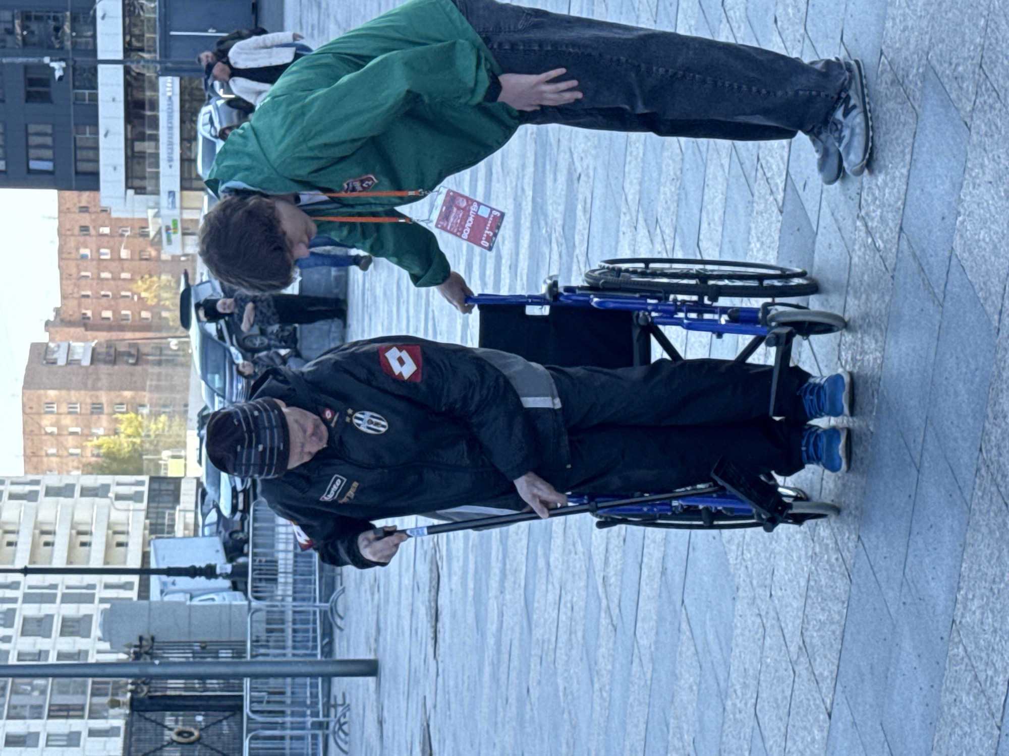 A man in a black jacket with the Juventus logo and a black and gray beanie is pushing a person in a wheelchair on a paved surface. The person being pushed is wearing a green jacket, jeans, and gray sneakers. A "Volunteer" lanyard is visible on the person in the green jacket. The background includes city buildings and several cars and people on the street. The photo was taken during the day in Yekaterinburg, Russia.