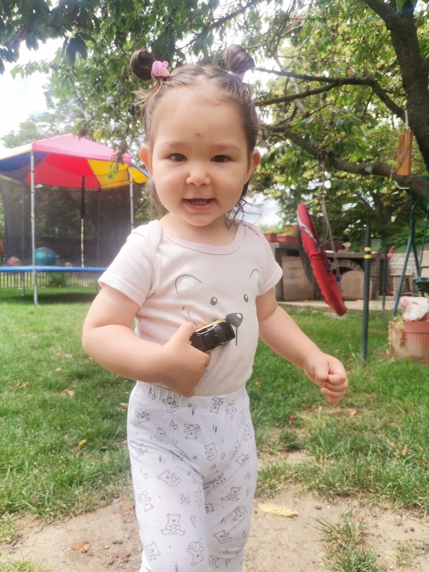 In the city of Osoitsa, Bulgaria, a young child, possibly of Caucasian and Asian descent, stands outdoors in what appears to be a backyard. The child, with light brown hair styled in two buns, wears a light pink t-shirt with a bear design and white leggings adorned with small teddy bear patterns. The child is holding a dark-colored toy in their right hand, near their chest. The background features a trampoline with a colorful canopy, a swing set, trees, and a grassy area. The lighting suggests it is daytime.
