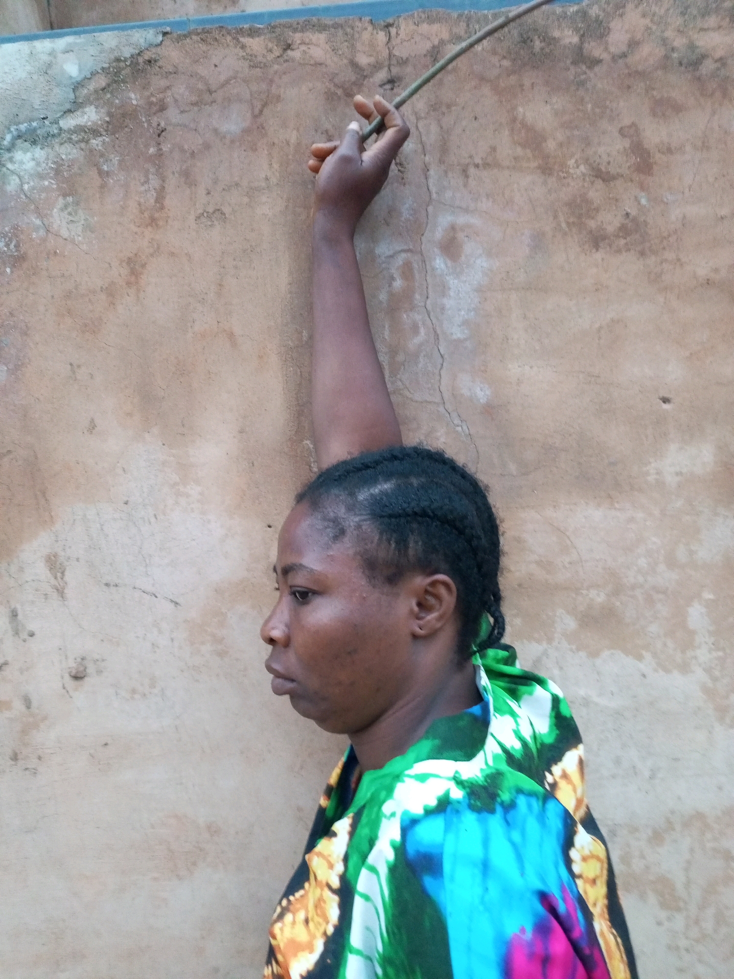 A medium shot, taken outdoors, shows a woman with dark skin and braided hair standing against a textured, light brown wall. The woman is positioned in the right half of the frame, facing left. Her right arm is raised vertically, with her hand gripping a thin, dark stick that extends above her head and towards the top of the frame. She is wearing a vibrantly colored shirt with a pattern of green, blue, yellow, and pink. The wall behind her is rough and shows signs of wear and staining. The image appears to be taken in Tempure, Nigeria.