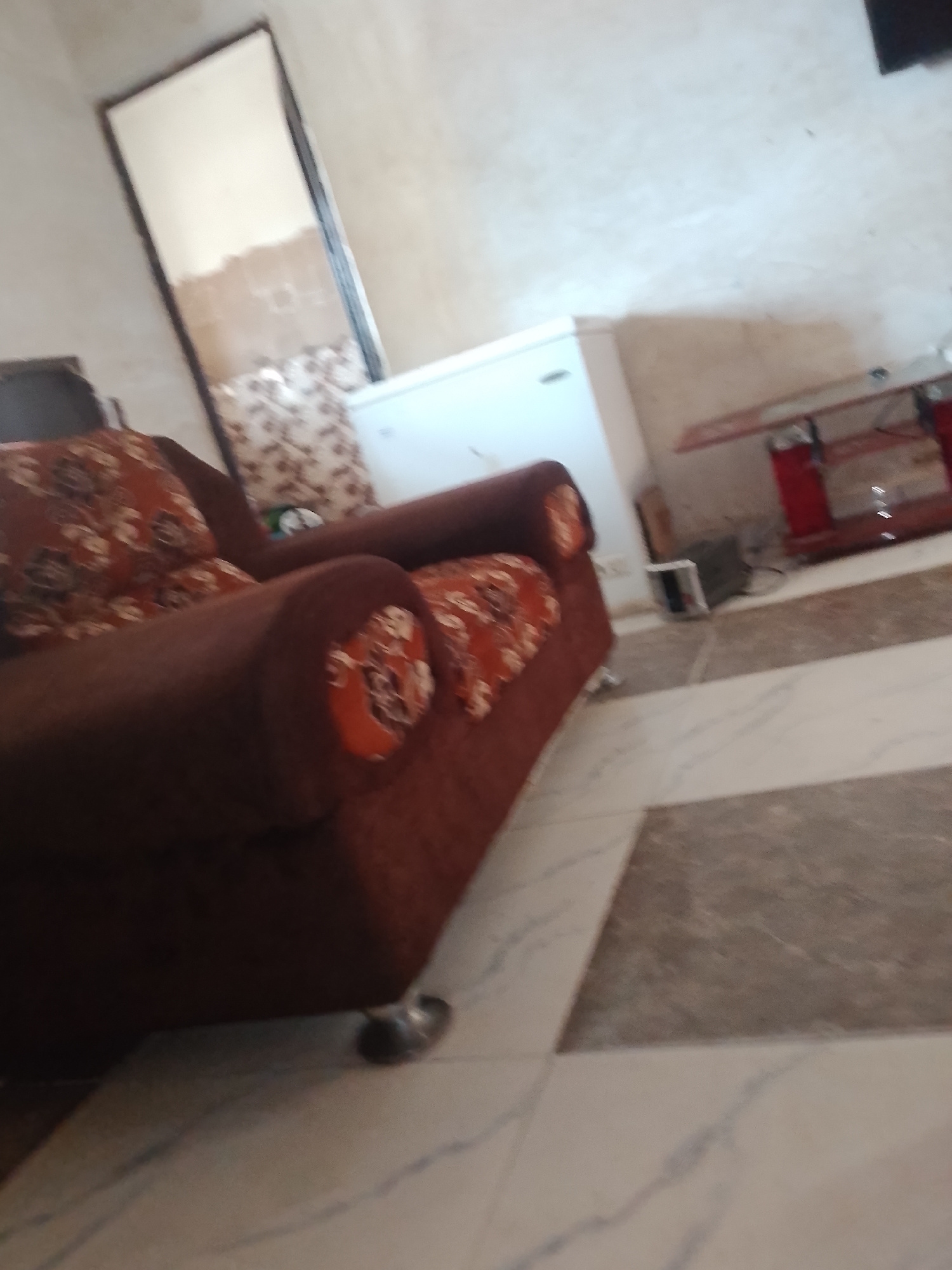 The image depicts an indoor scene, likely a living room or common area, with furniture and appliances visible. A large, dark brown sofa with patterned cushions is positioned in the foreground, appearing to be the primary subject of the photo. To the left of the sofa, a tall, rectangular mirror with a dark frame leans against the wall. In the background, a white refrigerator or similar appliance stands. Further to the right, a red entertainment center or shelf unit is visible, with some electronic devices on it. The floor is tiled with a pattern of light and dark marble-like squares. The overall lighting suggests an indoor setting, possibly during the daytime given the illumination. No people are present in the image, and no text is visible. The environment appears to be a residential or communal space in Jalingo, Nigeria.