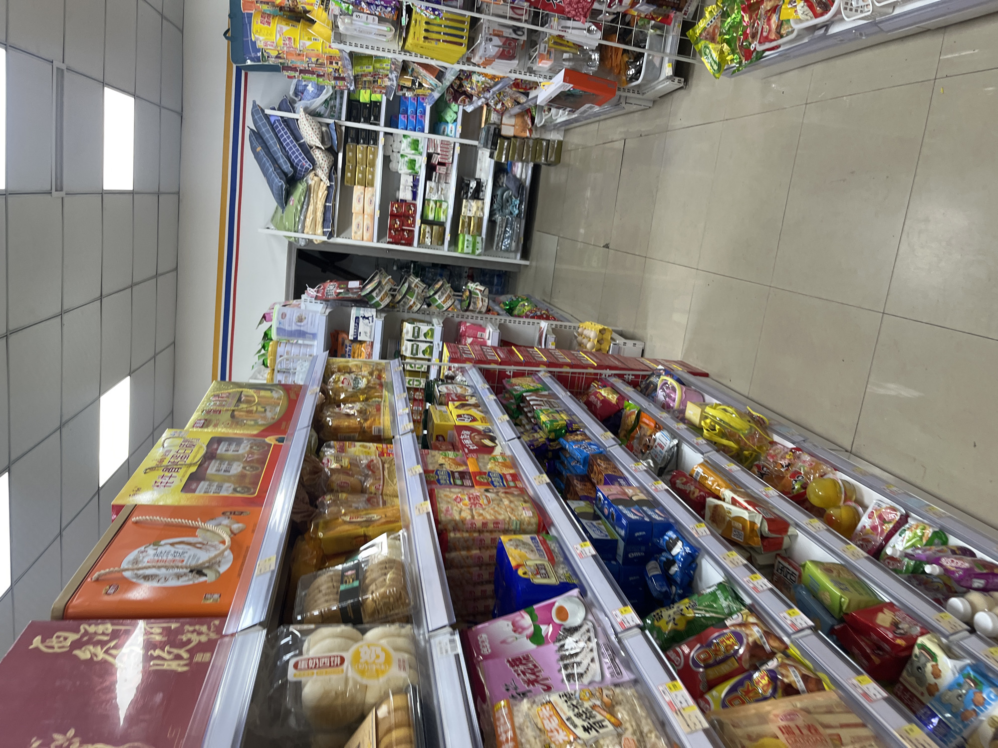 This is a photograph of a convenience store aisle in Yuzhong District, China. The shelves are stocked with various food items, including packaged snacks, drinks, and what appear to be pastries or bread. The aisles are clean and well-lit with fluorescent ceiling lights. The overall impression is one of a typical retail environment.