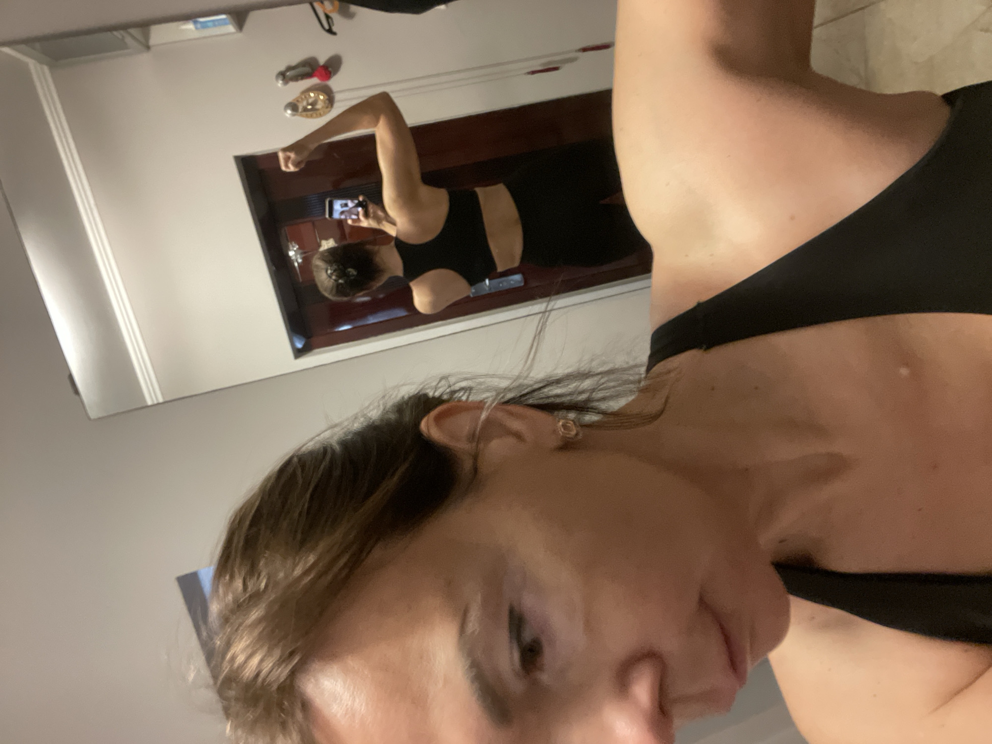 A woman is taking a selfie in a mirror, showing her flexing her bicep. She is wearing a black sports bra and black leggings. The background is a room with a mirror, a door, and some hooks on the wall. The floor is tiled. The lighting is artificial and warm. The location is unknown.