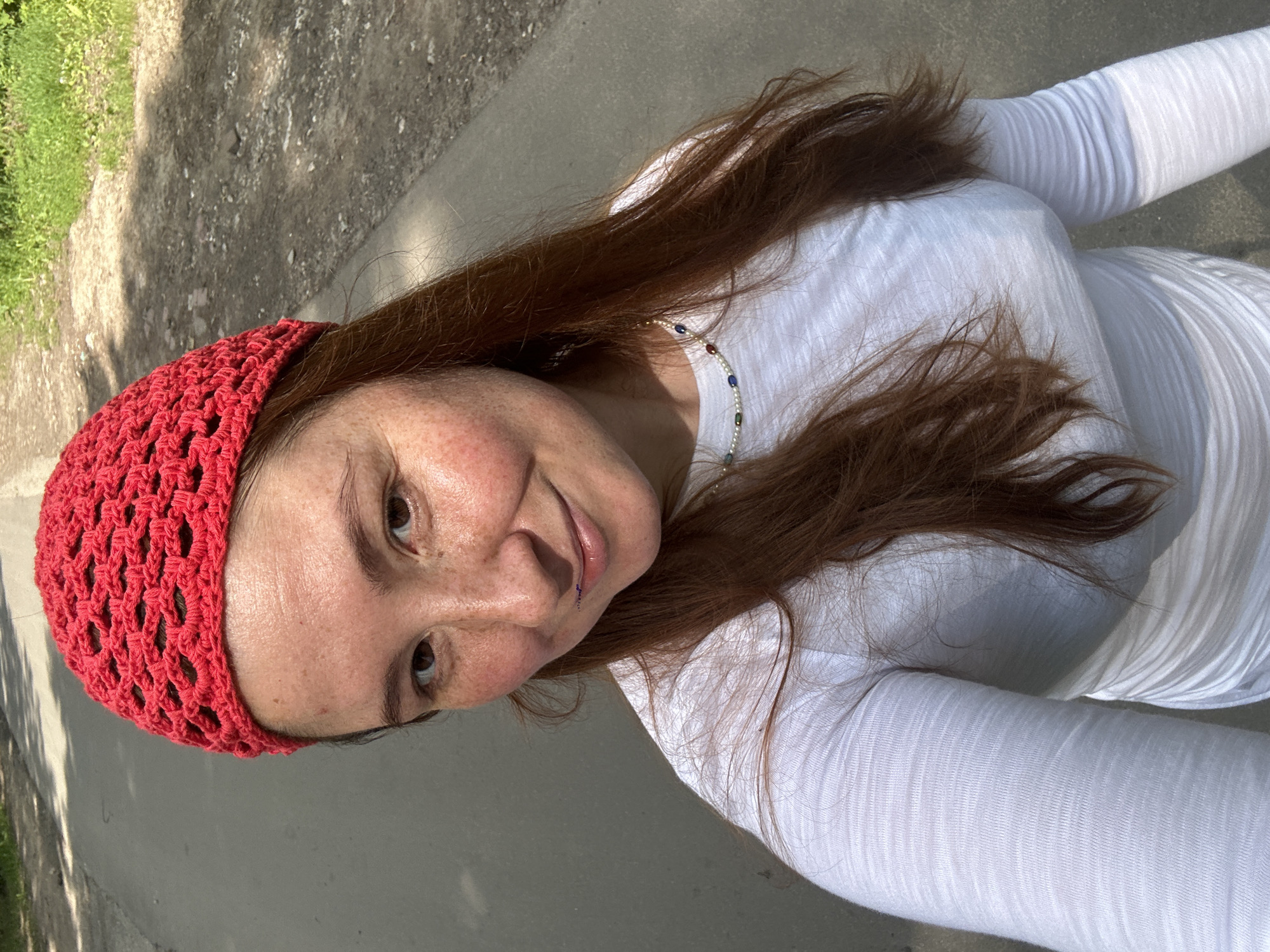 A fair-skinned woman with red hair and freckles wears a red knitted cap and a white long-sleeved shirt. She is outdoors, with a blurred background of a paved road and grassy areas suggesting a park or a road outside of a city. Her face is turned towards the camera with a slight smile. A beaded necklace is visible around her neck. The image appears to be a selfie, taken on a sunny day.