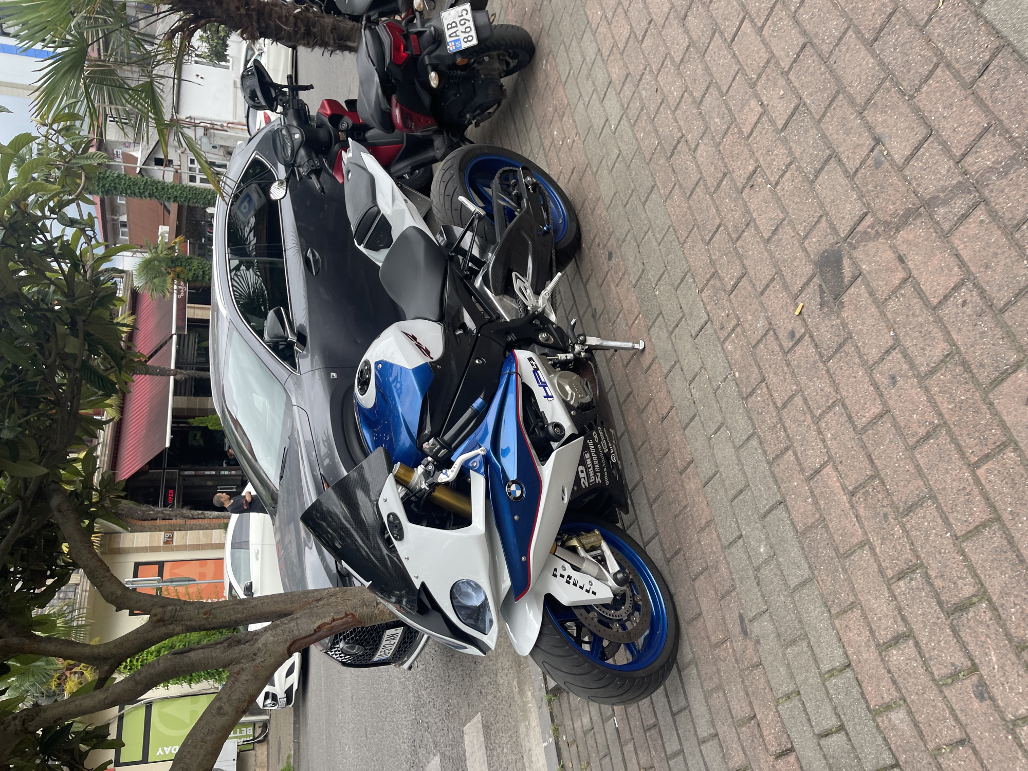 The image shows a street scene in Batumi, Georgia. The primary subjects are two motorcycles parked on a brick sidewalk next to a dark-colored car. In the foreground, a white, blue, and black sport motorcycle is prominently displayed, its front wheel and part of the fairing visible. Behind it, a red and black motorcycle is partially visible. A person can be seen leaning against the car in the background, looking away from the camera.
The scene appears to be an outdoor setting on a sidewalk. The background shows buildings with various architectural styles, including some with red awnings and balconies. A tree with thick branches partially obscures the view of the buildings and the car. The time of day is unclear, but the lighting suggests it could be daytime. The weather appears to be clear, with no visible rain or heavy clouds.
Notable details include the license plate on the red motorcycle, which appears to read "AB 8695". The sport motorcycle has "Pirelli" written on its front fender and various decals on its fairing. The car next to the motorcycles is a modern sedan. The presence of parked motorcycles and cars suggests this is a public street or parking area. The overall atmosphere seems calm and ordinary, capturing a moment of urban life.