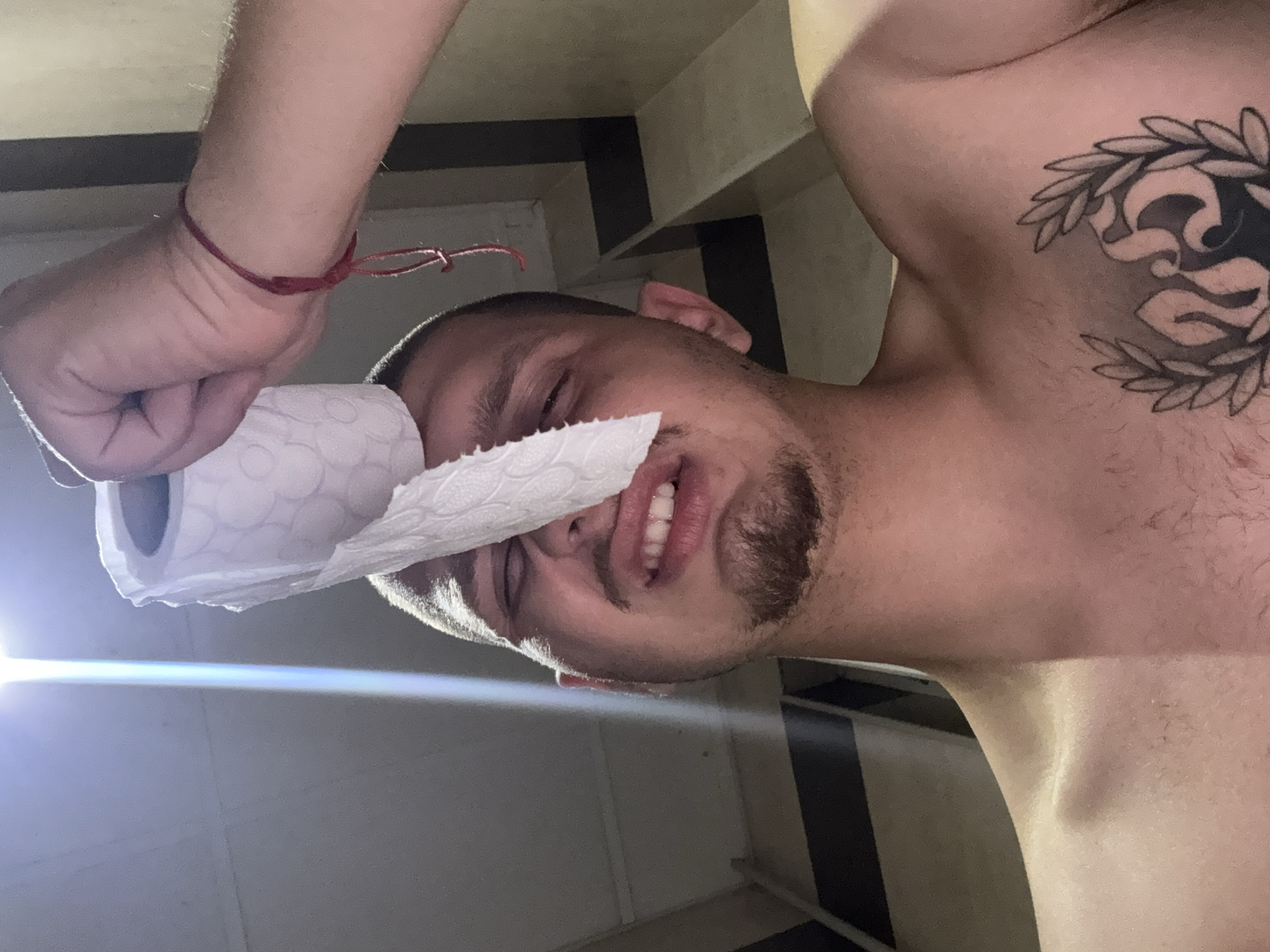 A low-angle, close-up shot shows a shirtless young man in Blagoevgrad, Bulgaria, with a roll of toilet paper held in front of his face, partially obscuring his nose and eyes. He has short dark hair, a goatee, and a tattoo on his chest. A bright light source shines from above and to the left, casting dramatic shadows. The background consists of textured walls with dark horizontal stripes, suggesting an indoor setting like a bathroom or hallway. A red string bracelet is visible on the wrist of a person's hand holding the toilet paper. The man is looking towards the camera with his mouth slightly open, revealing his teeth.
