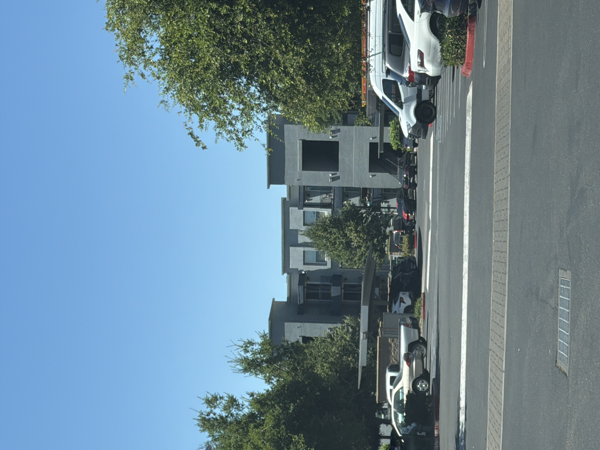 The image shows a parking lot in front of a modern, multi-story apartment building with a gray facade, visible under a clear blue sky. Several vehicles are parked in the lot, including a white van, a dark SUV, and a white sedan. Some greenery, including trees and bushes, is interspersed with the buildings and parking lot. The scene appears to be in a developed urban or suburban area, consistent with a location in Elk Grove, United States of America.