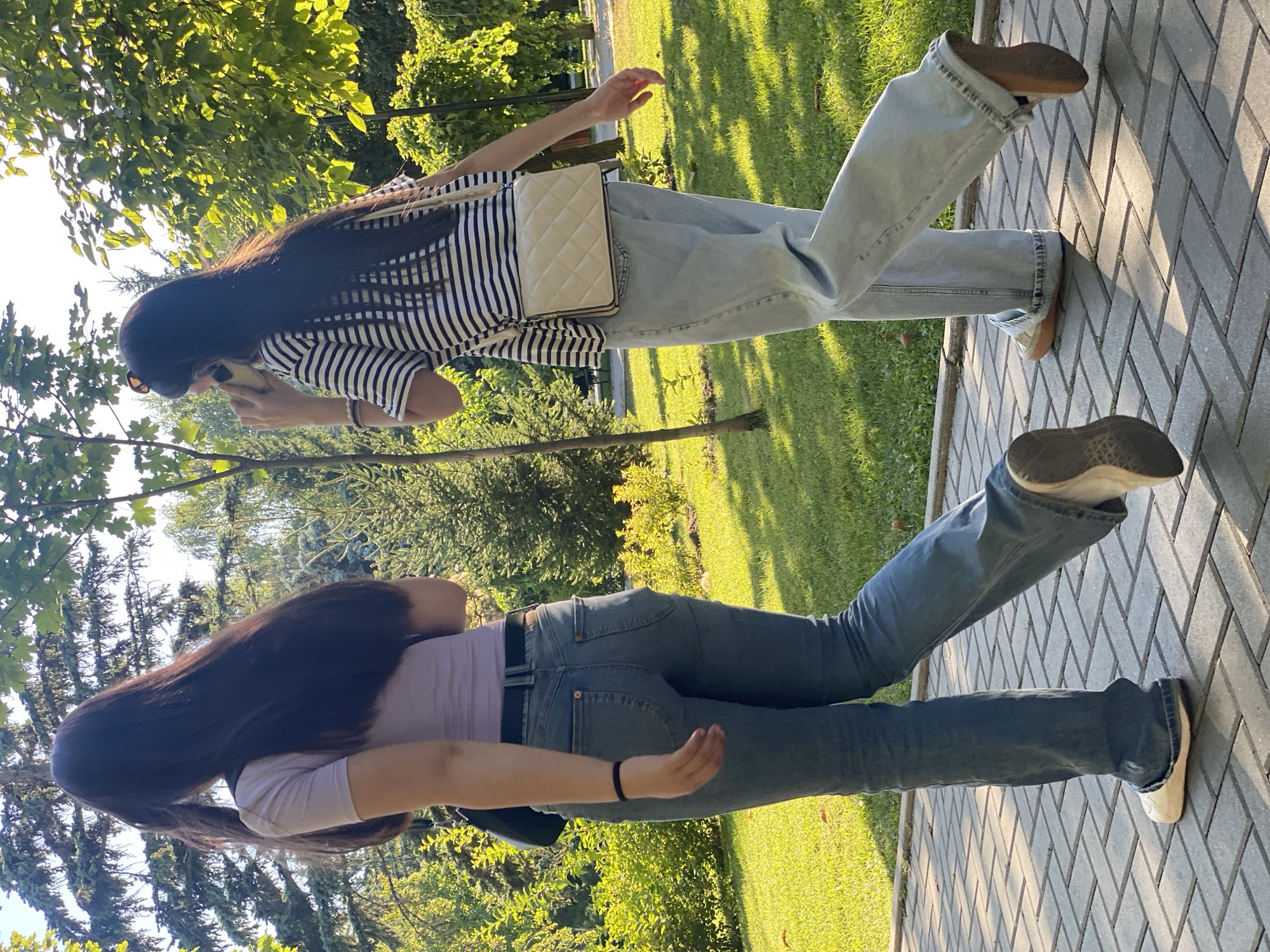 Two young women walk on a paved pathway in a park on a sunny day. The woman on the left, with long dark hair, wears a lavender t-shirt and blue jeans, carrying a dark bag over her shoulder. The woman on the right, also with long dark hair, wears a black and white striped t-shirt and light blue jeans, holding a cell phone to her ear and carrying a quilted beige crossbody bag. Both women are walking away from the camera, towards a grassy area with trees and bushes. The pathway is made of grey rectangular pavers arranged in a herringbone pattern. The sunlight casts distinct shadows on the pathway, indicating a low sun angle. The environment appears to be a well-maintained public park with lush green grass and trees, suggesting a natural setting within an urban area. The architecture or specific landmarks that would definitively place this scene in Chișinău, Moldova are not present in the image.