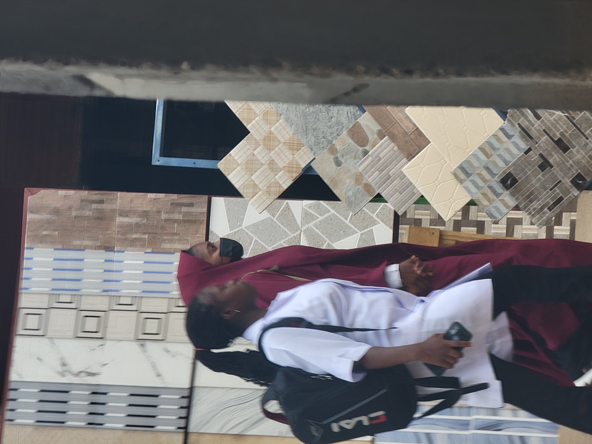 Two individuals are lying on a selection of tiles. One individual is wearing a white shirt and a black backpack. The other individual is wearing a maroon garment and a black face mask. The location is implied to be Maiburiji, Nigeria, based on the prompt.