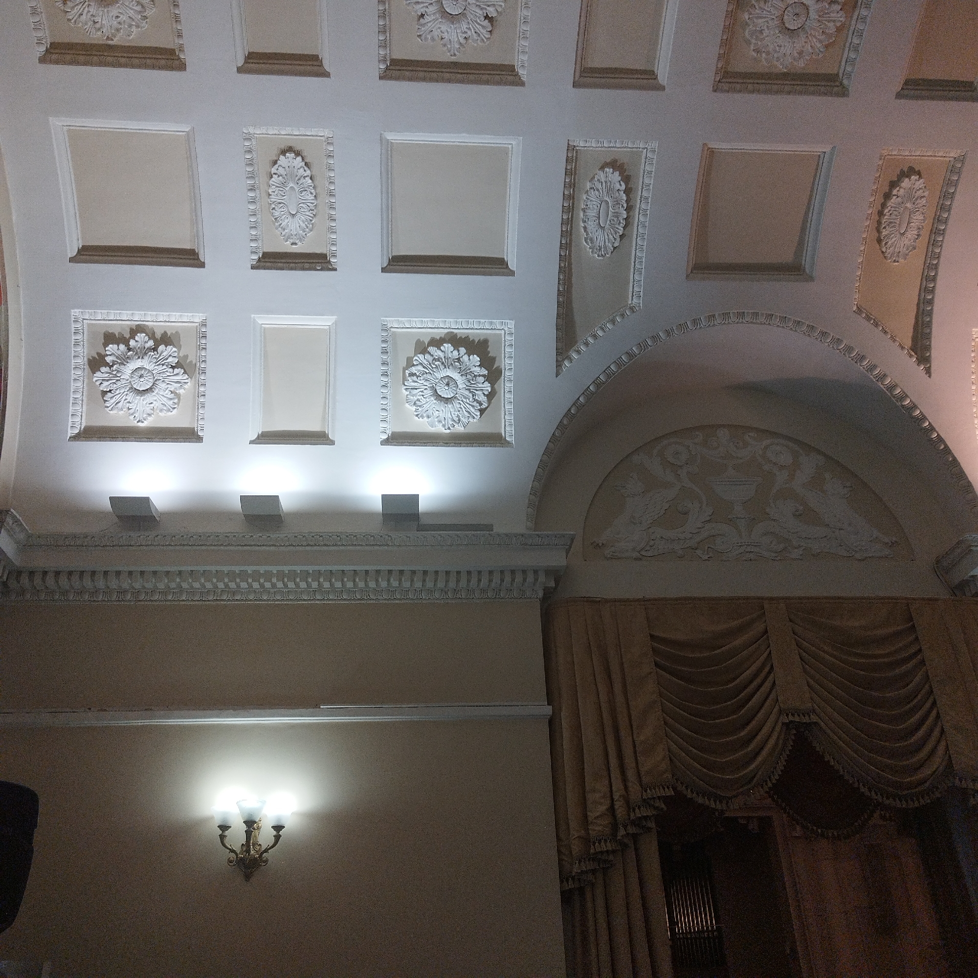 The image captures a section of the interior of a building in Yekaterinburg, Russia, focusing on architectural details. The ceiling is adorned with a grid of square and rectangular panels, some plain and others featuring ornate floral rosettes. Beneath the ceiling, a decorative frieze runs along the top of the wall, punctuated by regularly spaced rectangular blocks. Below the frieze, a wall sconce with three bulbs provides illumination, casting light on the wall. To the right, a large archway frames draped curtains. Inside the arch, a sculpted relief with a central vase flanked by stylized figures is visible. The overall impression is of a formal, possibly historical, interior space.