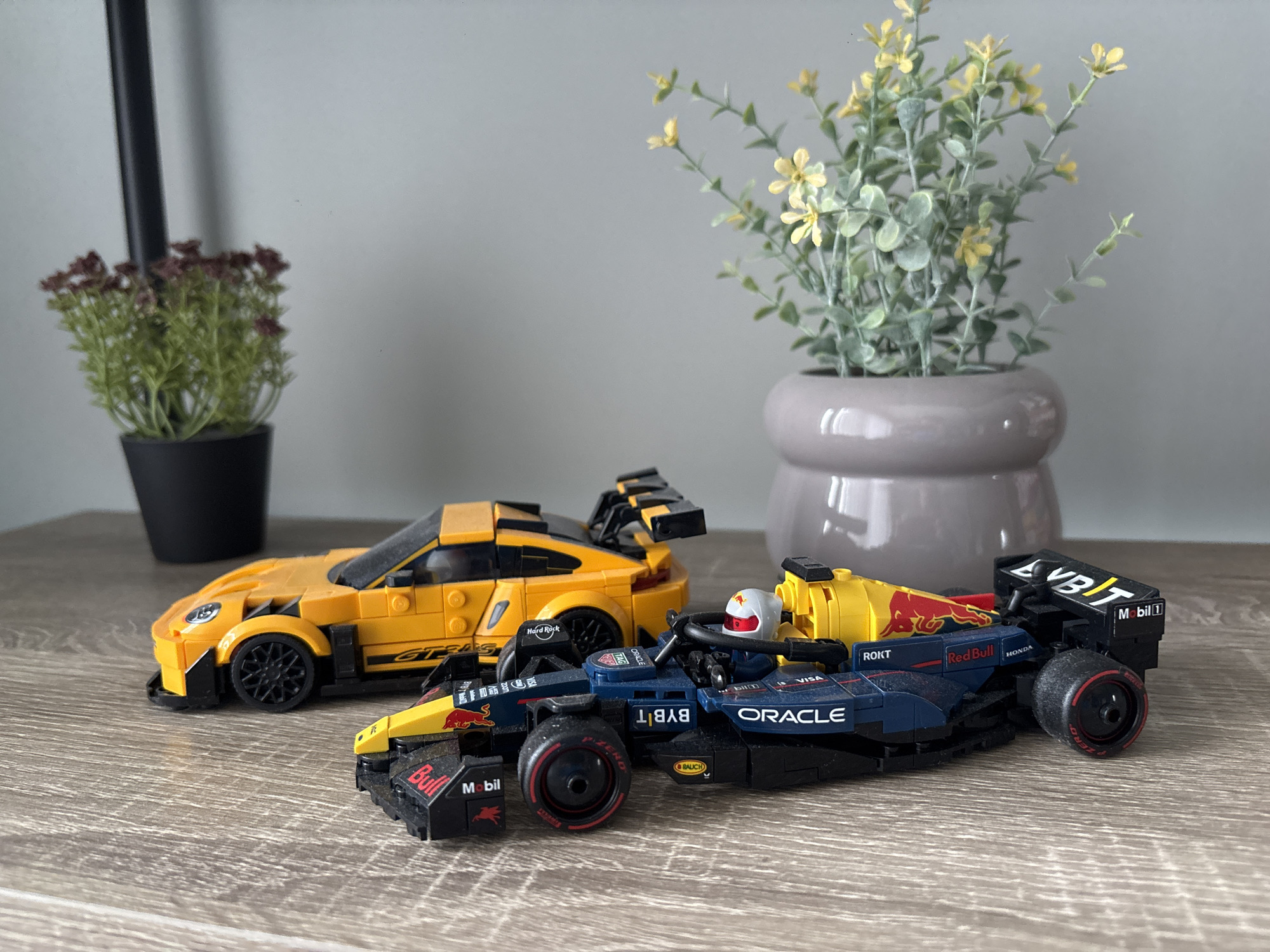 The image shows two Lego race cars positioned on a wood-grained surface. The first car is yellow with black wheels and a black spoiler, and bears the logo "Hard Rock" and "GTRA5". The second car is predominantly navy blue and red, with markings such as "Oracle", "Red Bull", "Mobil", and "BYBIT". The car appears to have a Lego figure seated in the cockpit wearing a white helmet. Behind the cars, there is a potted plant with yellow flowers in a gray pot, and another potted plant with reddish-brown flowers in a black pot. The background is a plain gray wall. The setting is likely indoors.