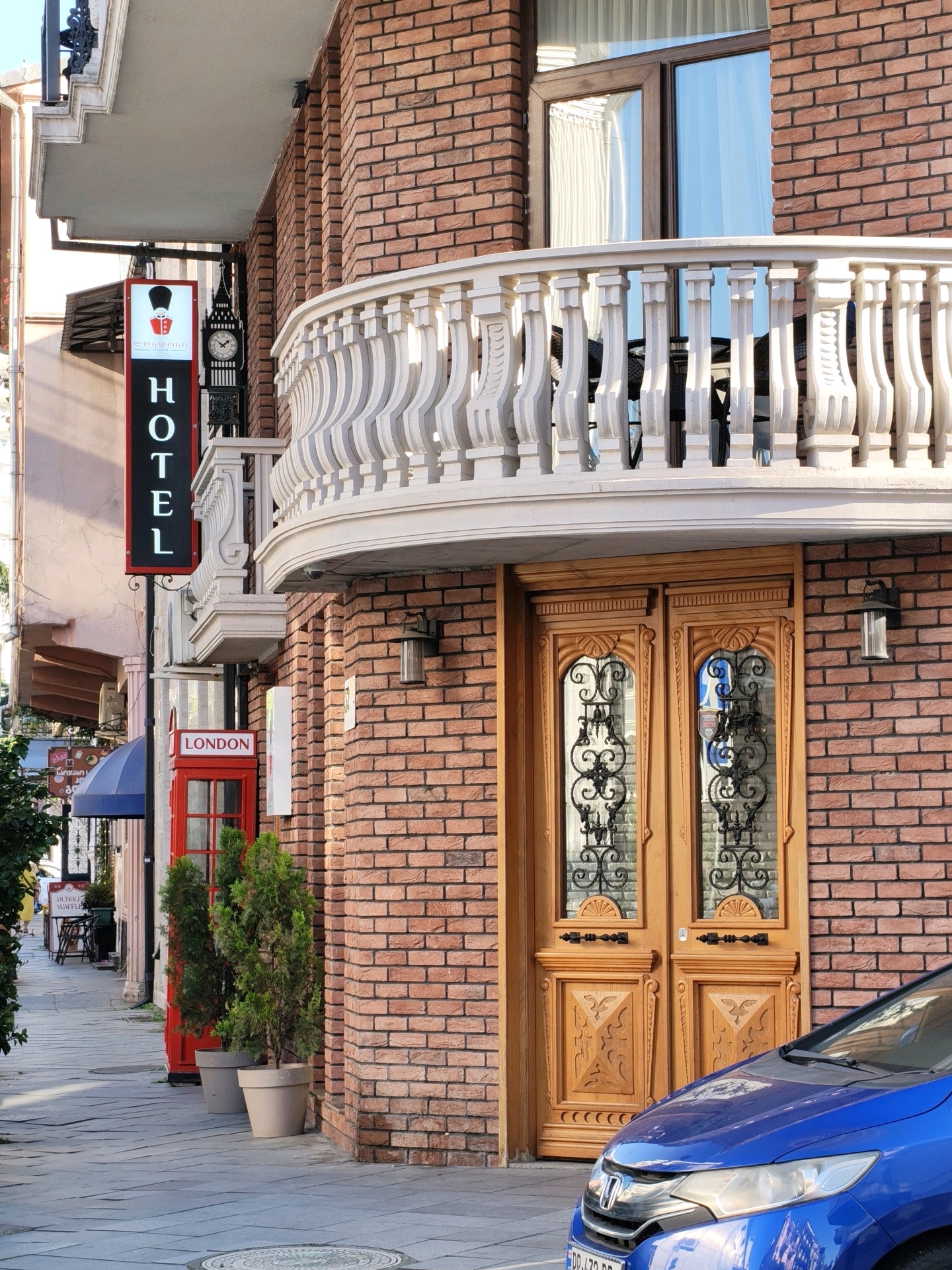 An exterior view of a hotel entrance on a street in Batumi, Georgia, features a red brick building with a decorative white balcony and ornate wooden doors. A vertical sign reading "HOTEL" in white letters on a black background is mounted on the building's facade. To the left, a red London-style phone booth with a "LONDON" sign is visible. Two potted evergreen trees stand in front of the doors. A blue car is parked in front of the entrance. The street is paved with grey stone tiles, and a blue awning is extended from an adjacent building. In the background, several signs and storefronts are visible, suggesting a commercial area. The lighting indicates it is daytime.