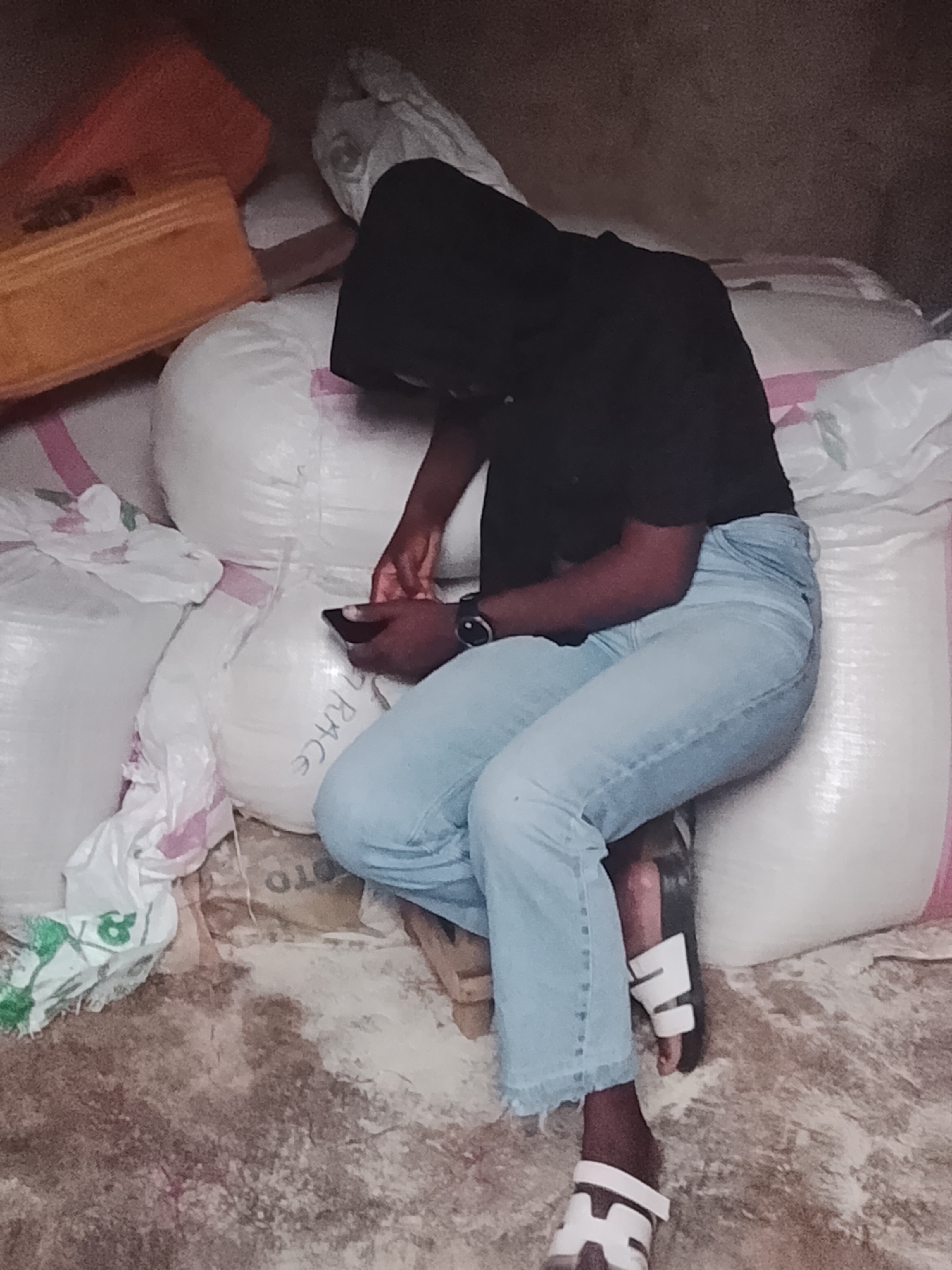 A person wearing a black hooded sweatshirt and light blue jeans sits on a pile of large white bags, possibly sacks of rice as one is labeled "RICE," holding a phone in their hands. The location appears to be indoors, possibly a storage area, in Jos, Nigeria. The floor is dirty. The person is also wearing sandals and a watch. There is an orange container atop the bags.