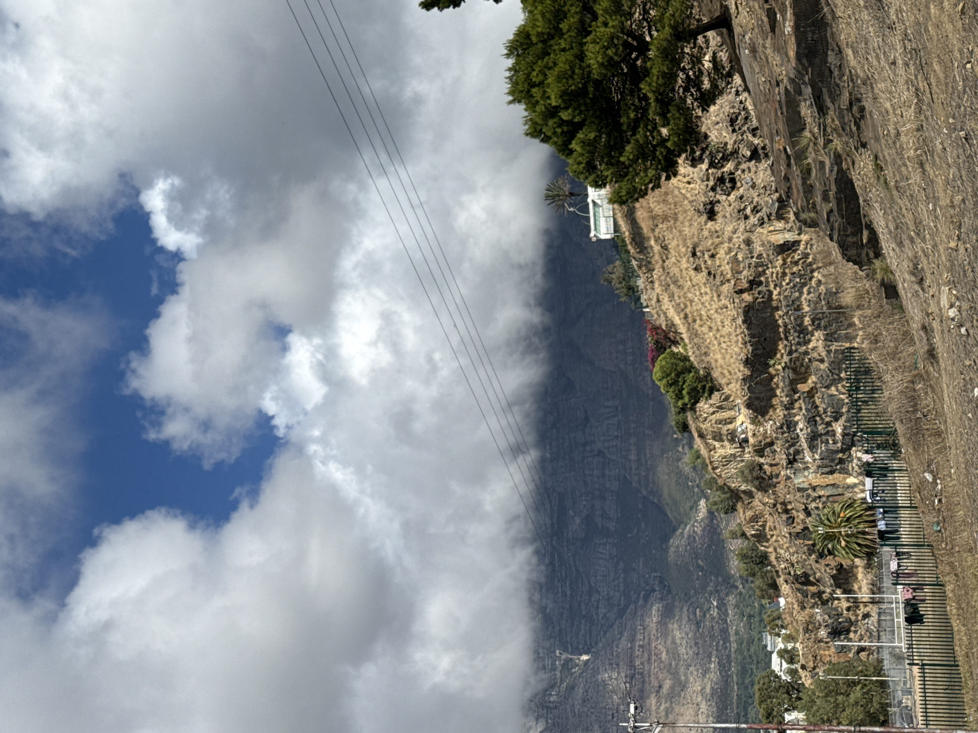 The image shows Table Mountain in Cape Town, South Africa, partially obscured by clouds. A cliff face with vegetation runs along the right side of the image. Power lines are visible in the upper left. Buildings can be seen near the base of the mountain.