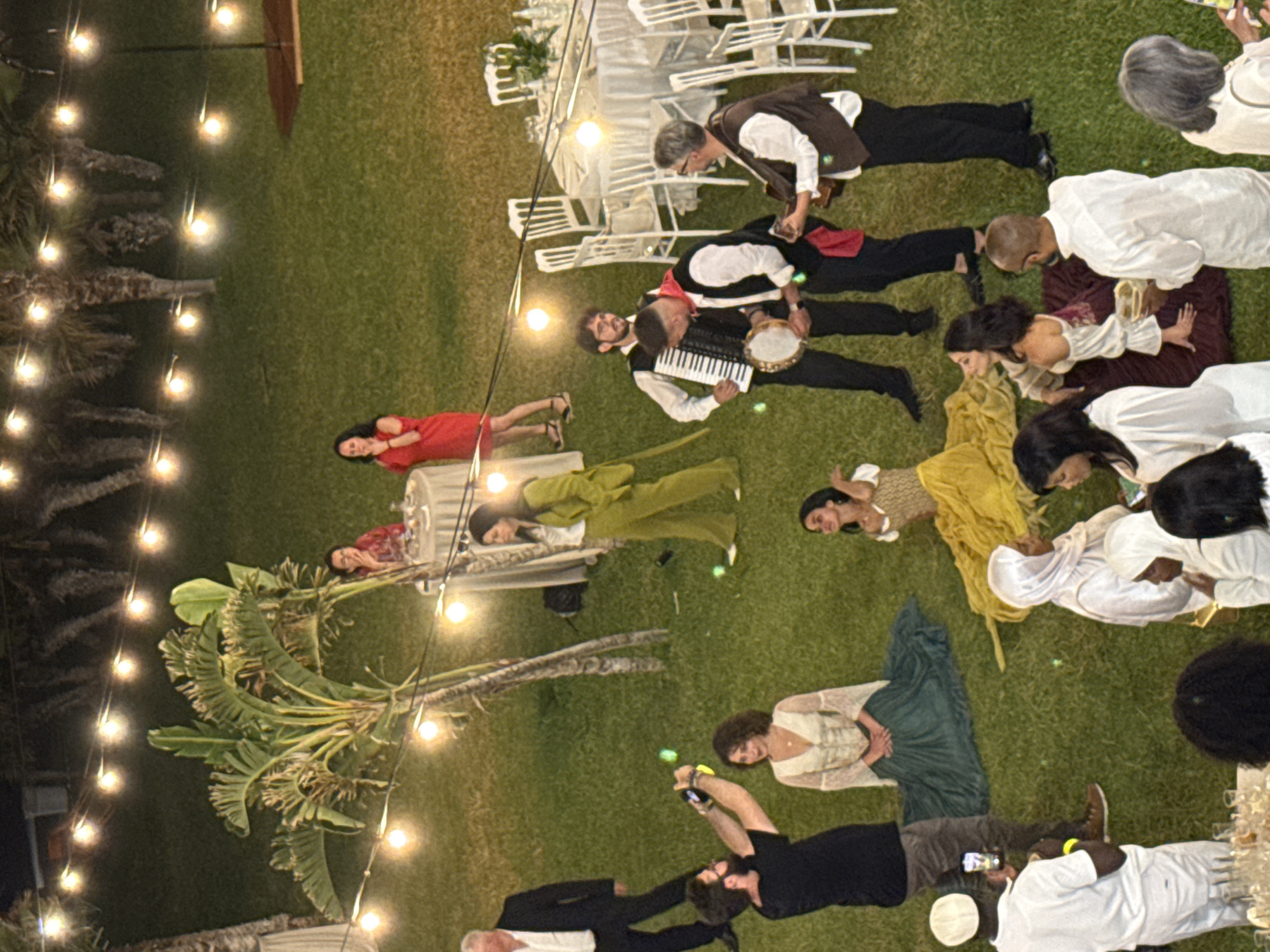 The image shows a nighttime outdoor scene, likely a staged performance or event, on a grassy area in Isola delle Femmine, Italy. String lights illuminate the area, which includes several individuals in various costumes. Some are lying on the grass, while others stand or kneel. There are elements of musical performance, with one person playing an accordion and another a tambourine. Several white chairs are arranged in the background, possibly for an audience or part of a set. The lighting suggests a casual, potentially theatrical atmosphere.