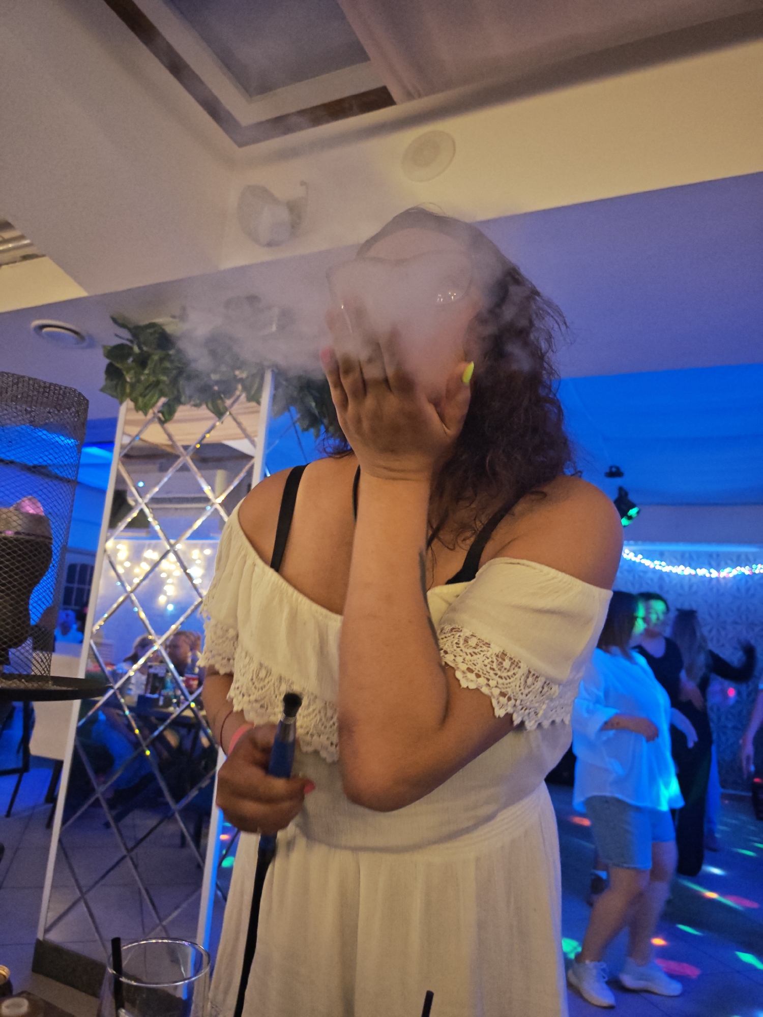 A woman stands in a room filled with blue light and other people. She wears a white dress with black straps and holds a hookah pipe. Smoke obscures her face. In the background, people stand and dance under colored disco lights, and there is a mirrored wall with decorative lights. There is a hookah on the left. The city name and location are not possible to verify from the image content.