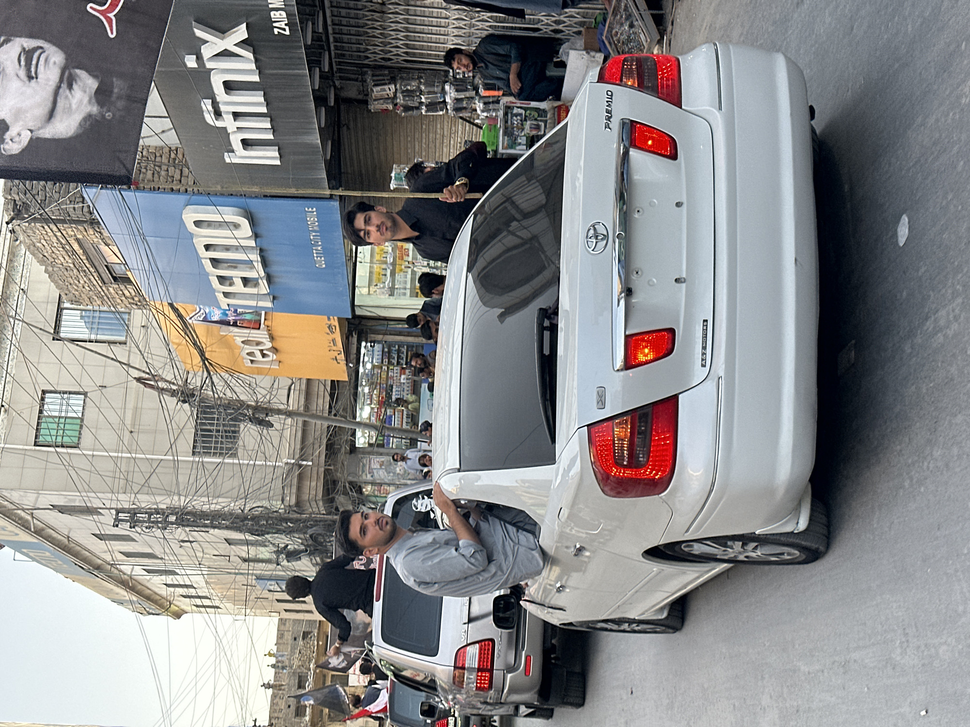 In a street scene in Unknown, Unknown, a procession of cars is observed moving down a street lined with buildings. The focus is on a white Toyota Premio car, with its rear facing the camera, displaying red taillights. A young man, wearing a grey shirt, is leaning out of the driver's side window, looking towards the right. Behind this car, a silver SUV is visible, with a person in a black shirt standing on its roof. Further back, another person in a black shirt is seated in the front passenger seat of the SUV, holding a long stick. The buildings lining the street feature large signs, including "TECNO" in blue and "INFINIX" in black. The overall atmosphere suggests a public gathering or event taking place during daylight hours.