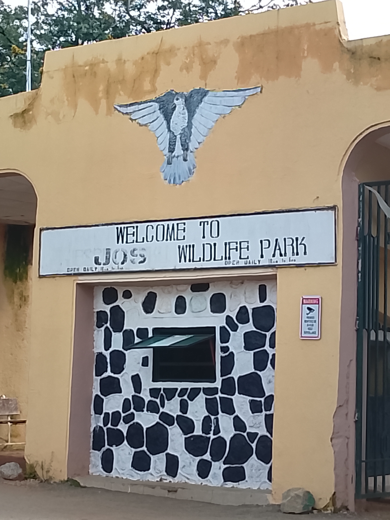The image depicts the entrance to Jos Wildlife Park in Nigeria. The facade of the building is painted a pale yellow, with a mural of a bird with outstretched wings above the entrance. A white sign with black lettering reads "WELCOME TO JOSS WILDLIFE PARK" and below that "OPEN DAILY 10 AM TO 6 PM". The entrance itself features a rough, stone-like pattern of black and white. A small window with a green awning is visible in the stone facade. To the right of the entrance, there is a red and white warning sign indicating surveillance. A metal gate can be seen to the far right of the image. The environment suggests an outdoor setting with trees visible in the background and a dirt or gravel path in the foreground.