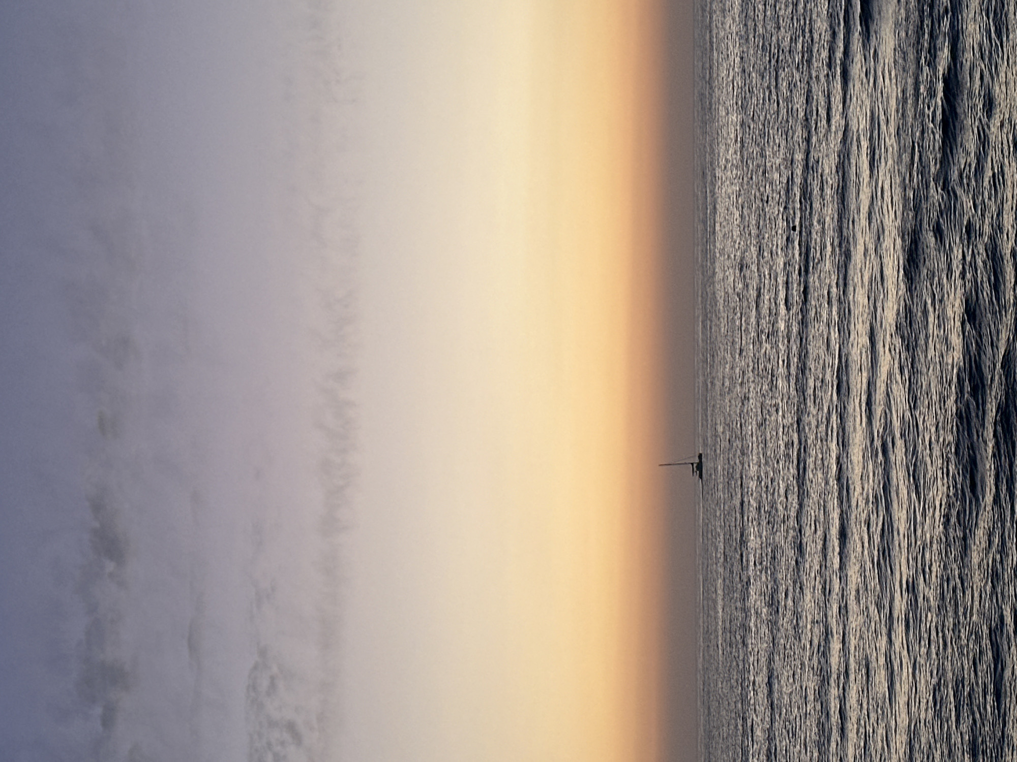 A photograph of the sea at sunrise or sunset. The sky is a gradient of blue-gray, light beige, and orange. On the right side is the sea with white-tipped waves. A sailboat is in the middle of the photograph, in the sea. The image appears to be taken in Tel Aviv, Israel.