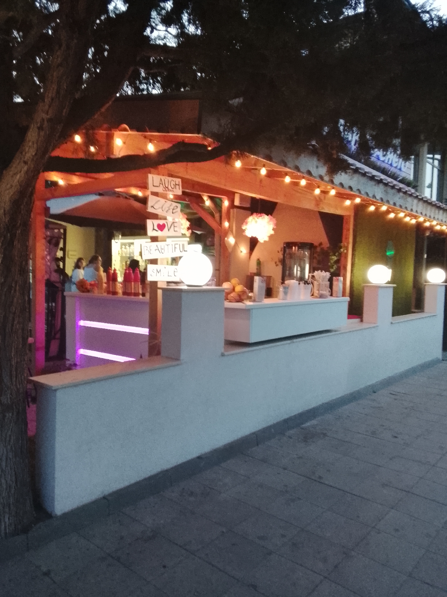 The image shows an outdoor bar in Nesebar, Bulgaria. The bar is decorated with string lights and has a wooden roof. Signs on the bar read "LAUGH", "Life", "LOVE", "BEAUTIFUL", and "SMILE". There are several people visible behind the bar, and bottles and glasses are on display. The bar is lit with pink and purple lights, and two large white spherical lamps are placed outside the bar. The ground in front of the bar is paved with grey tiles. A large tree is to the left of the bar. The overall scene suggests a vibrant nightlife or entertainment venue.