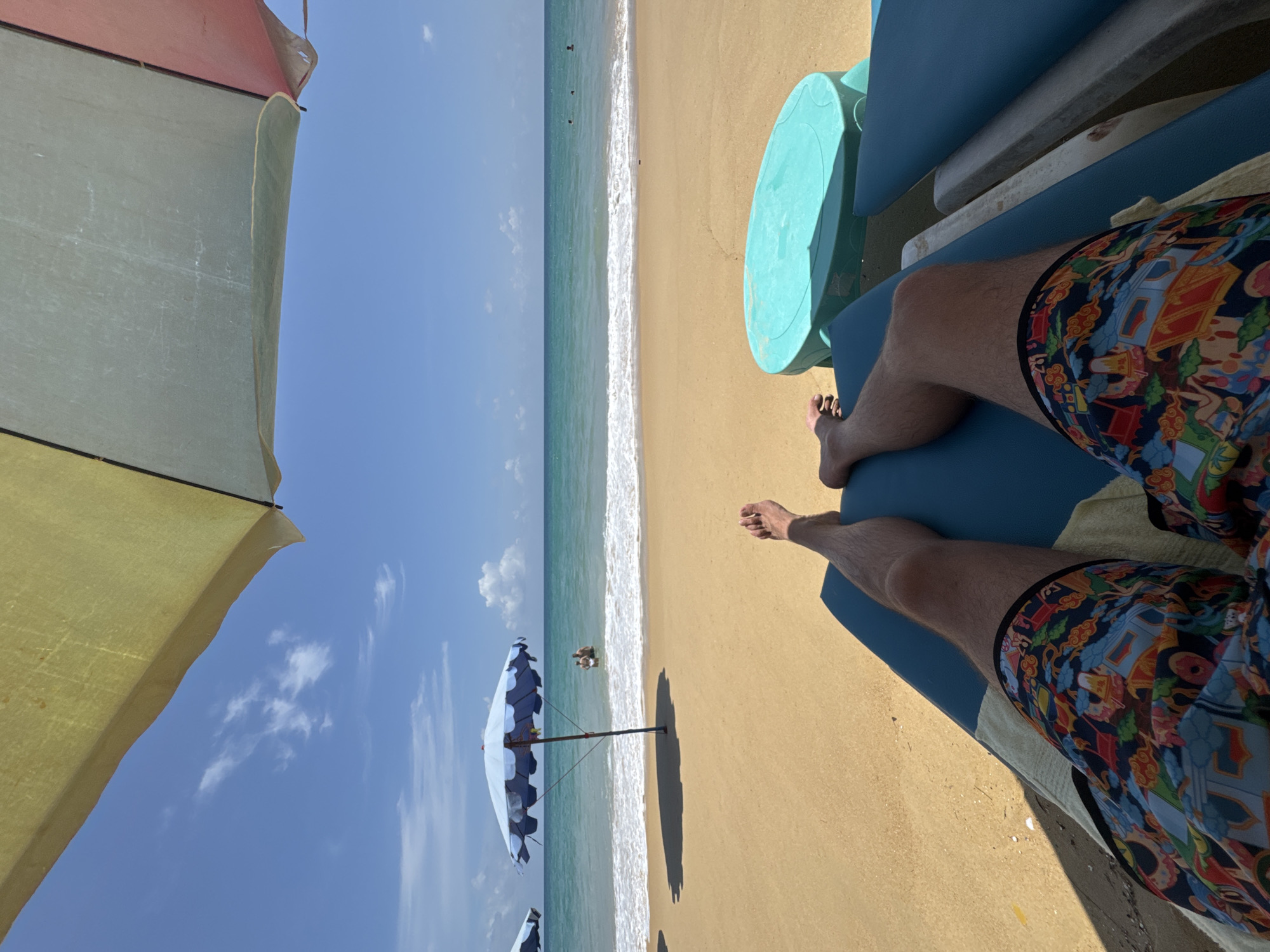 The image shows a first-person perspective of a person relaxing on a lounge chair on Karon Beach, Thailand. The person is wearing colorful patterned shorts and their legs are visible. In the background, the turquoise ocean meets the sandy beach, with gentle waves rolling in. Two beach umbrellas are visible, one blue and white, and another with yellow, green, and pink panels, providing shade. Several other people are in the water in the distance. A teal-colored plastic table is positioned on a lounge chair to the right of the frame. The sky is a clear blue with a few white clouds, and the sun is shining brightly.