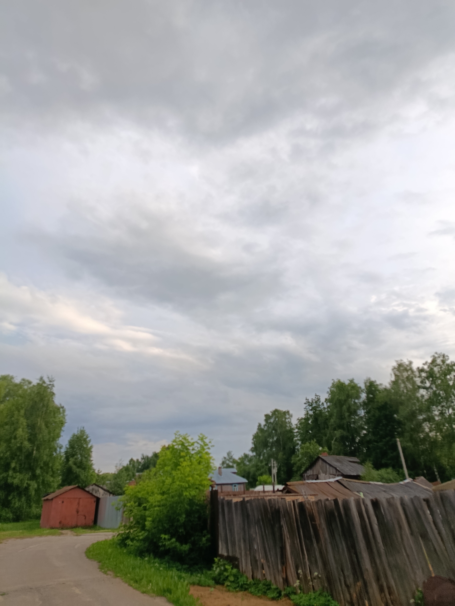 The image captures a scene in the city of Vereya, Russia. A winding paved road curves through a sparsely populated area. On the left side of the road, a weathered red metal garage stands, with another metal structure adjacent to it. Lush green trees and bushes dominate the landscape. In the background, more trees are visible, along with several rustic wooden structures and a fence constructed from weathered wooden planks. The sky is overcast with a thick layer of gray clouds.