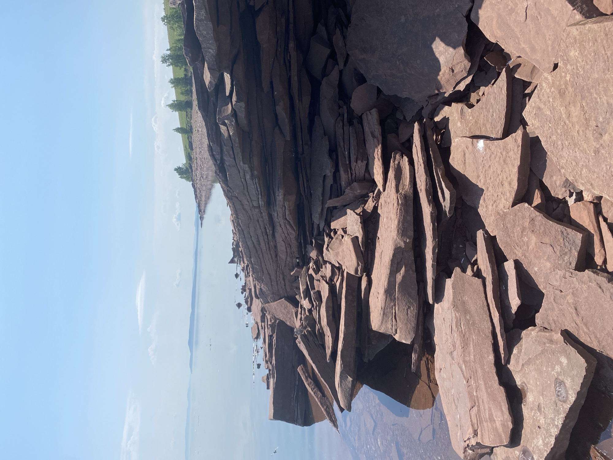 The image depicts a rocky shoreline on a clear, sunny day. The foreground is dominated by large, flat, reddish-brown rocks, some of which are broken into smaller pieces. The rocks are piled and scattered, with some extending into the calm, clear water. In the middle ground, the shoreline curves away, lined with more of these flat rocks and a strip of green grass with scattered trees. The background shows a vast expanse of calm water, with distant, hazy landmasses or islands on the horizon under a light blue sky with a few scattered white clouds. The overall impression is one of a natural, undeveloped waterfront. The image does not contain any people or provide any specific clues about its location within Ust'-Erba, Russia, or any other city.