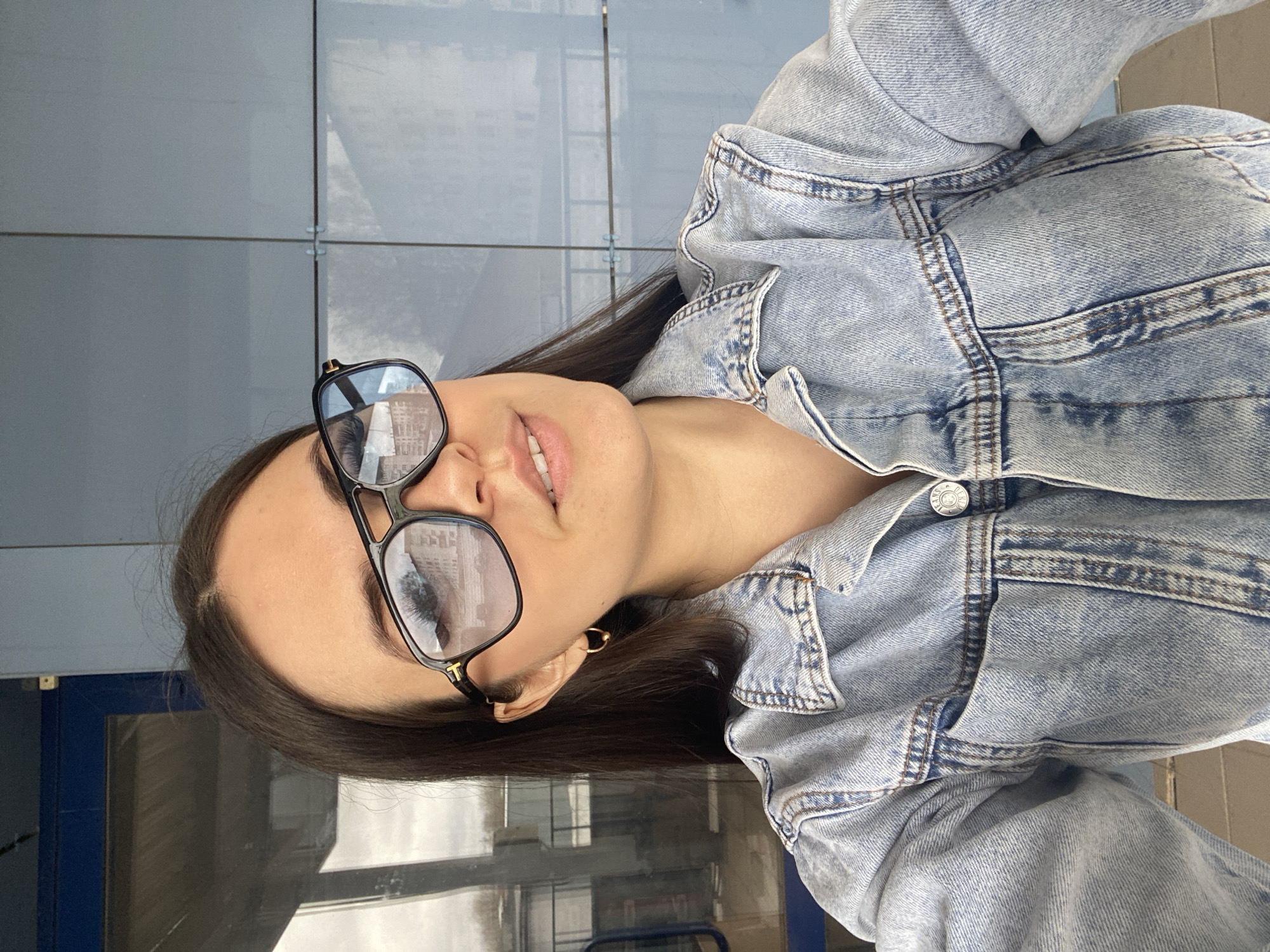 A woman with dark hair and sunglasses is wearing a light-wash denim jacket. The location is the city of Chaikovsky, Russia. The background is composed of square blue and grey tiles that may be a building facade. The woman is positioned at an angle to the camera.