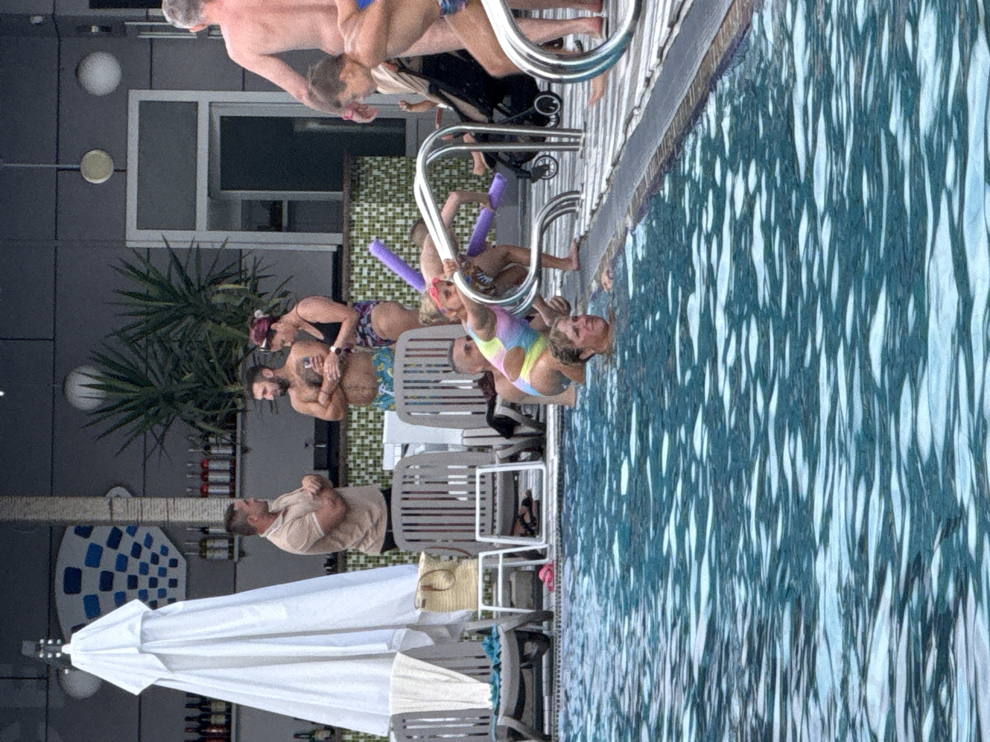 A group of people are gathered around a swimming pool. In the foreground, a portion of the pool's shimmering blue water is visible. In the mid-ground, several individuals are in or near the water. A woman with blonde hair is partially submerged, looking to her left. To her right, a young girl with blonde hair, wearing goggles and a multi-colored swimsuit, holds onto a purple pool noodle and leans on a silver railing. Behind them, an adult woman with dark hair and a dark swimsuit is holding a phone. Further back, a man with a beard is shirtless, holding what appears to be a drink. Another man in a tan shirt is talking to him. Behind them, a bar area is visible with bottles on shelves. Several lounge chairs are lined up near the pool, with a large white umbrella set up nearby. A baby stroller is visible to the right of the pool railing. The background shows a building with a tiled facade and some foliage. The scene appears to be an outdoor resort or hotel area.