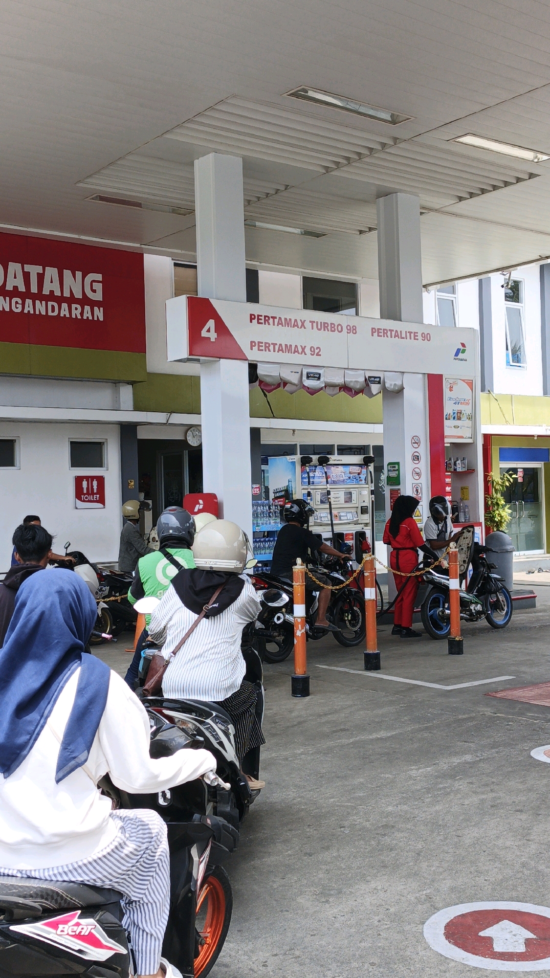 The image depicts a queue of people on motorcycles at a Pertamina gas station in Pangandaran, Indonesia. The gas station has a white canopy supported by white columns. A large red sign with white text, partially obscured, reads "DATANG NGENDARAAN" which likely signifies a vehicle entrance or welcome. Above the gas pumps, a white sign displays "4" in a red circle, followed by the names of fuel types: "PERTAMAX TURBO 98", "PERTALITE 90", and "PERTAMAX 92".
Several individuals are present, predominantly on motorcycles. In the foreground, a person wearing a blue hijab and a white shirt with blue and white striped pants sits on a black and pink motorcycle. Behind them, another person in a white and black striped shirt and black pants is seated on a motorcycle. To the right, a person in a red uniform, presumably a gas station attendant, is interacting with someone on a motorcycle. Further back, other people on motorcycles and attending to the pumps are visible. The ground is paved concrete with red and white directional arrows. Various gas pumps are lined up, and some people are in the process of refueling. The overall scene suggests a busy day at a common service facility.