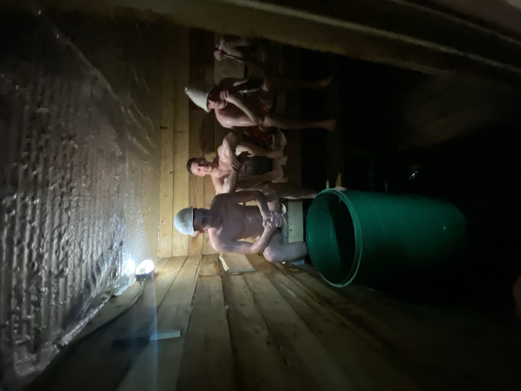 The image shows the interior of a sauna. Three shirtless men are visible inside, one wearing a white hard hat. The walls are constructed of wood paneling, and one wall is covered with a metallic-looking material. A green barrel is positioned on the floor near the bottom right of the image. A small light fixture is also visible near the bottom left. It is described to be in the city of Chaikovsky, Russia.