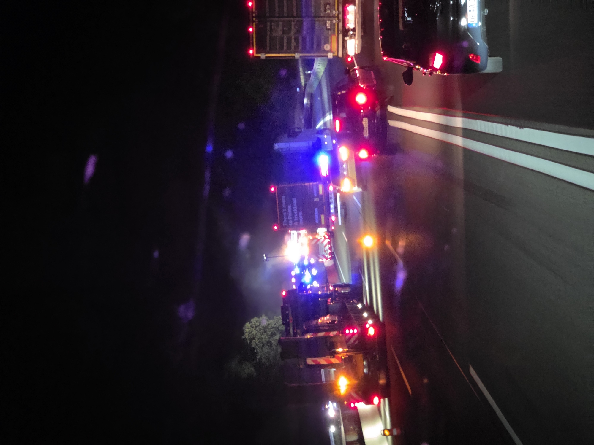 The image depicts a nighttime scene on a highway, likely in the vicinity of Seligenstadt, Germany, due to the license plate visible on a car. Several vehicles are present, including large trucks and a police car with flashing blue lights. One truck has a sign that reads "There's more to Prime. A truckload more." The road is well-lit by the vehicles' headlights and taillights, as well as by streetlights in the distance. The overall impression is one of a minor incident or a traffic stop. There is some light fog or mist in the air, causing reflections from the lights. The environment appears to be a multi-lane highway.