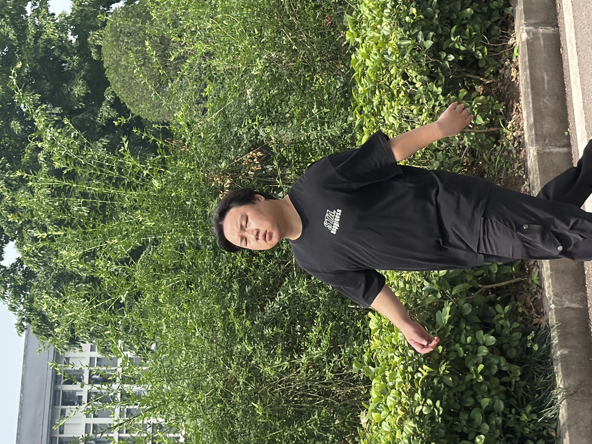 An outdoor, eye-level shot captures a person of East Asian descent standing amidst lush green foliage, likely in Jiangjin, China, on a sunny day. The person, appearing to be male, is dressed in a black short-sleeved t-shirt and black pants. His arms are slightly outstretched, with open palms facing forward, and his head is tilted slightly. His eyes are partially closed, suggesting he might be squinting due to the bright sun or wearing a contemplative expression. On his t-shirt, the words "STA." and "happiness" are visible. In the background, dense green bushes and trees are prominent, with a building featuring multiple windows partially visible on the left side. A concrete curb or sidewalk runs along the right edge of the frame, separating the green space from an unseen path or road. The bright lighting indicates it is daytime.